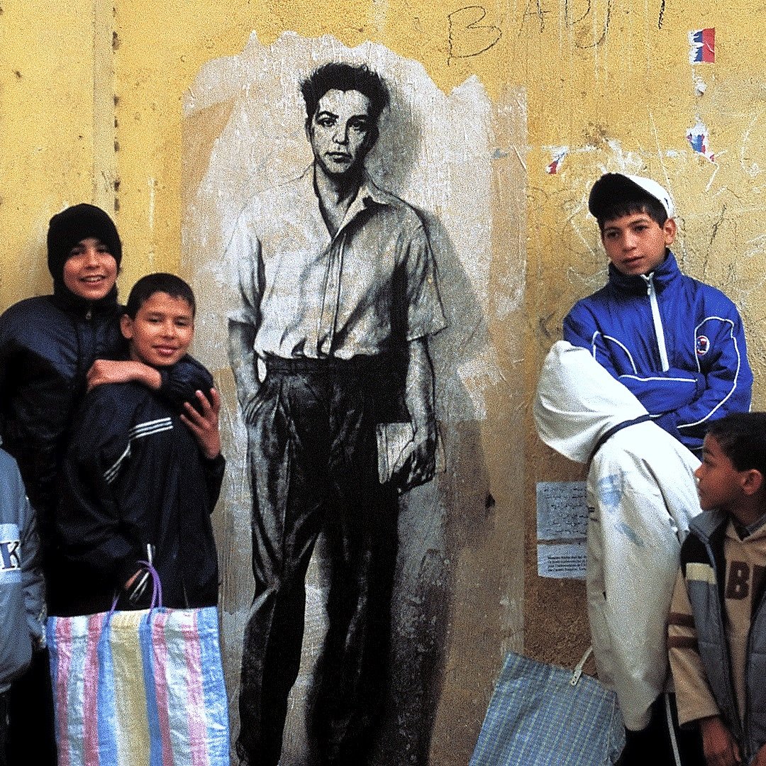 The image features a group of children standing in front of a large mural on a yellow wall. The mural depicts a black-and-white portrait of a young man in a shirt. The children are dressed in casual, colorful clothing, with some wearing hats. They appear to be interacting playfully, with a sense of camaraderie among them. The background shows some graffiti and markings, adding to the urban setting.