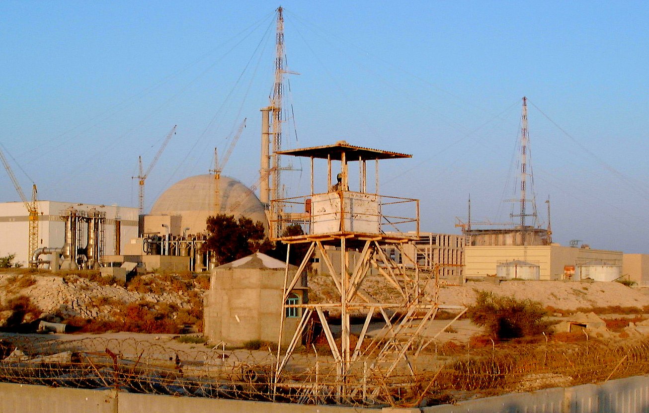 L'image présente un site industriel, probablement une centrale nucléaire, avec plusieurs structures. On peut voir une grande infrastructure circulaire au fond, entourée de diverses installations et grue. Au premier plan, il y a une tour de garde sur une plateforme surélevée, indiquant probablement un niveau de sécurité élevé dans cette zone. Des barbelés et des éléments de sécurité entourent l'endroit, suggérant une zone protégée. Le ciel est clair, ce qui donne une lumière naturelle à la scène.