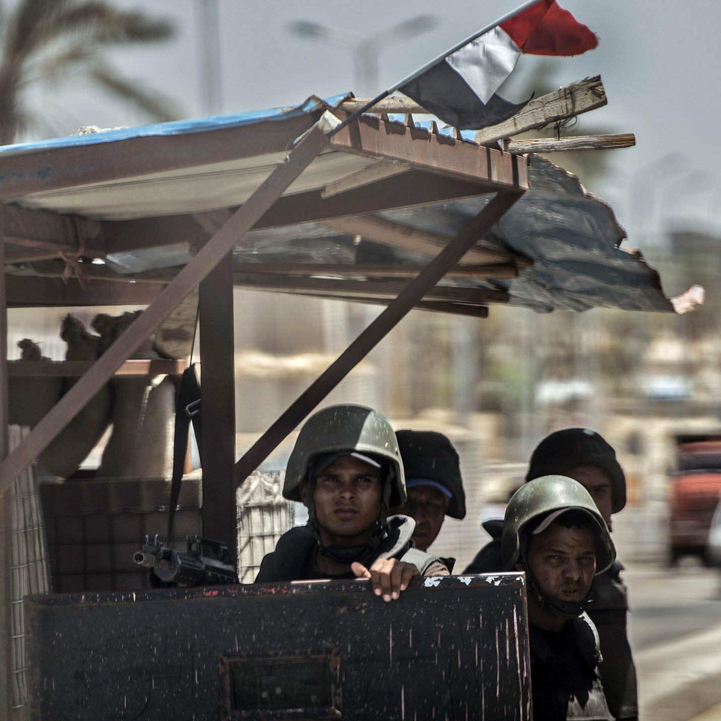 L'image montre un groupe de soldats, probablement en uniforme militaire, postés à un poste de contrôle. Ils semblent attentifs et vigilants, avec des casques de protection. On remarque également un drapeau qui flotte sur le poste, indiquant un contexte éventuellement national ou militaire. L'arrière-plan dépeint une rue, suggérant une ambiance urbaine, et on peut apercevoir des véhicules au loin. Le temps semble clair, mais la scène pourrait transmettre une sensation de tension ou de vigilance.