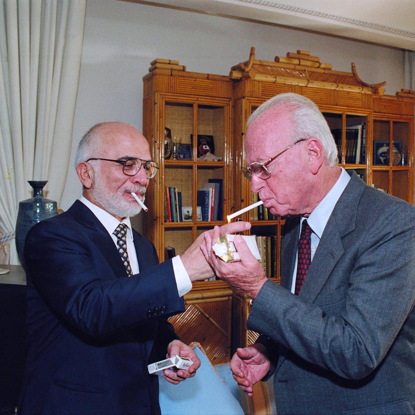 L'image montre deux hommes, chacun tenant une cigarette. L'un des hommes, vêtu d'un costume sombre et portant des lunettes, allume la cigarette de l'autre homme, qui porte également un costume et a une barbe grise. Ils se tiennent dans une pièce élégante, avec des meubles raffinés et des étagères en arrière-plan. L'atmosphère semble conviviale et intime.