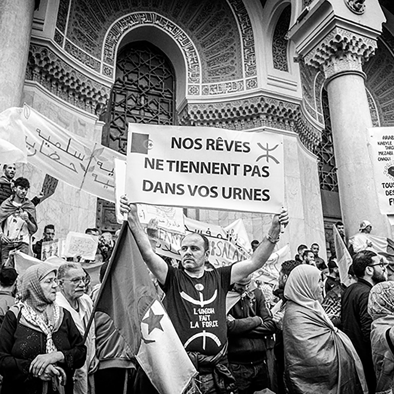 L'immagine ritrae una manifestazione di protesta. Al centro, un uomo tiene in alto un cartello con scritto "NOS RÊVES NE TIENNENT PAS DANS VOS URNES", che significa "I nostri sogni non stanno nelle vostre urne". Sullo sfondo, ci sono altri manifestanti e striscioni recanti vari messaggi. L'atmosfera sembra intensa, con persone che esprimono il loro dissenso e la loro determinazione. L'immagine è in bianco e nero, aggiungendo un senso di drammaticità e urgenza alla scena.