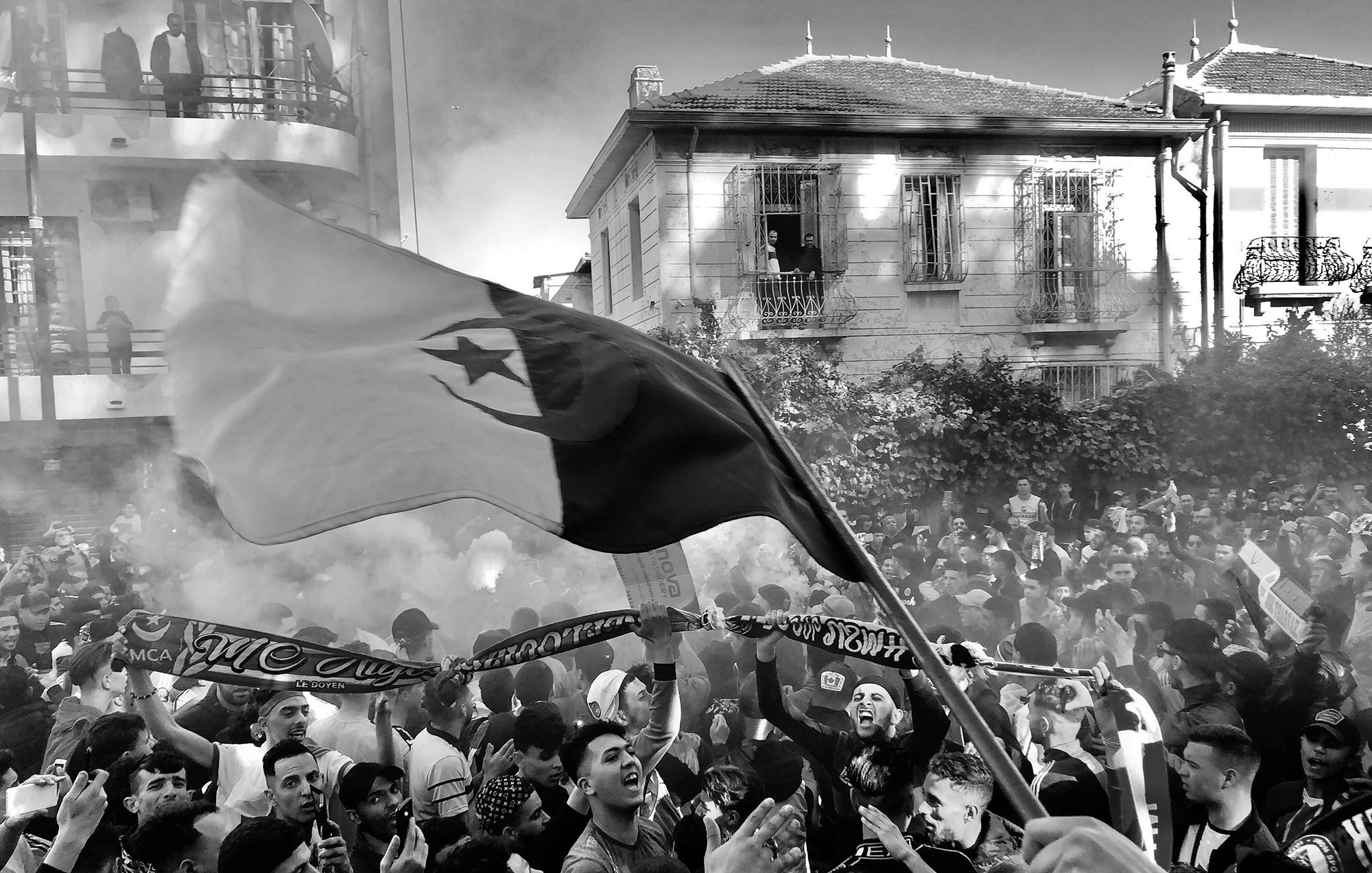 La imagen muestra a una gran multitud en un ambiente festivo, probablemente durante un evento deportivo o una celebración. Se pueden ver personas ondeando una bandera, así como pancartas y bufandas, lo que sugiere un fuerte sentido de identidad y apoyo a un equipo. El ambiente está lleno de humo, lo que añade un efecto dramático a la escena. La gente parece emocionada y unida, disfrutando del momento. Las edificaciones al fondo indican que la actividad está ocurriendo en un área urbana.