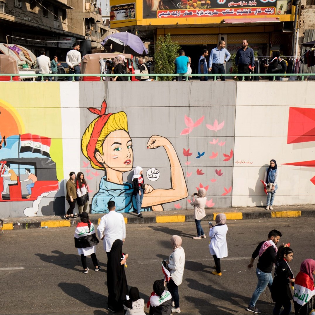 L'immagine mostra un murale su un grande muro divisorio, con diverse sezioni artistiche. A sinistra, c'è un ritratto di un uomo con una barba, che sembra essere ispirato a un personaggio storico o culturale, accompagnato da elementi grafici che richiamano il paesaggio urbano. A destra, c'è l'immagine di una donna in posa iconica, che rappresenta la forza e l'emancipazione, con dettagli colorati e dinamici. In basso, si osserva un gruppo di persone che passeggiano lungo la strada, creando un contrasto tra l'arte e la vita quotidiana. La scena riflette una vivace interazione tra arte, cultura e comunità.