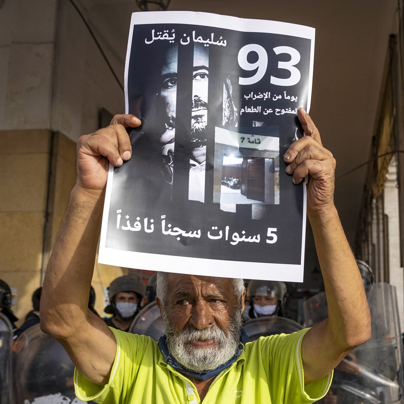 L'image montre un homme âgé tenant une pancarte en arabe. La pancarte mentionne des chiffres et des phrases qui semblent évoquer une question de justice ou de prison. En arrière-plan, on aperçoit des policiers en tenue d'émeute, ce qui suggère une atmosphère de protestation ou de tension. L'homme exprime probablement un sentiment d'injustice ou de revendication sociale.