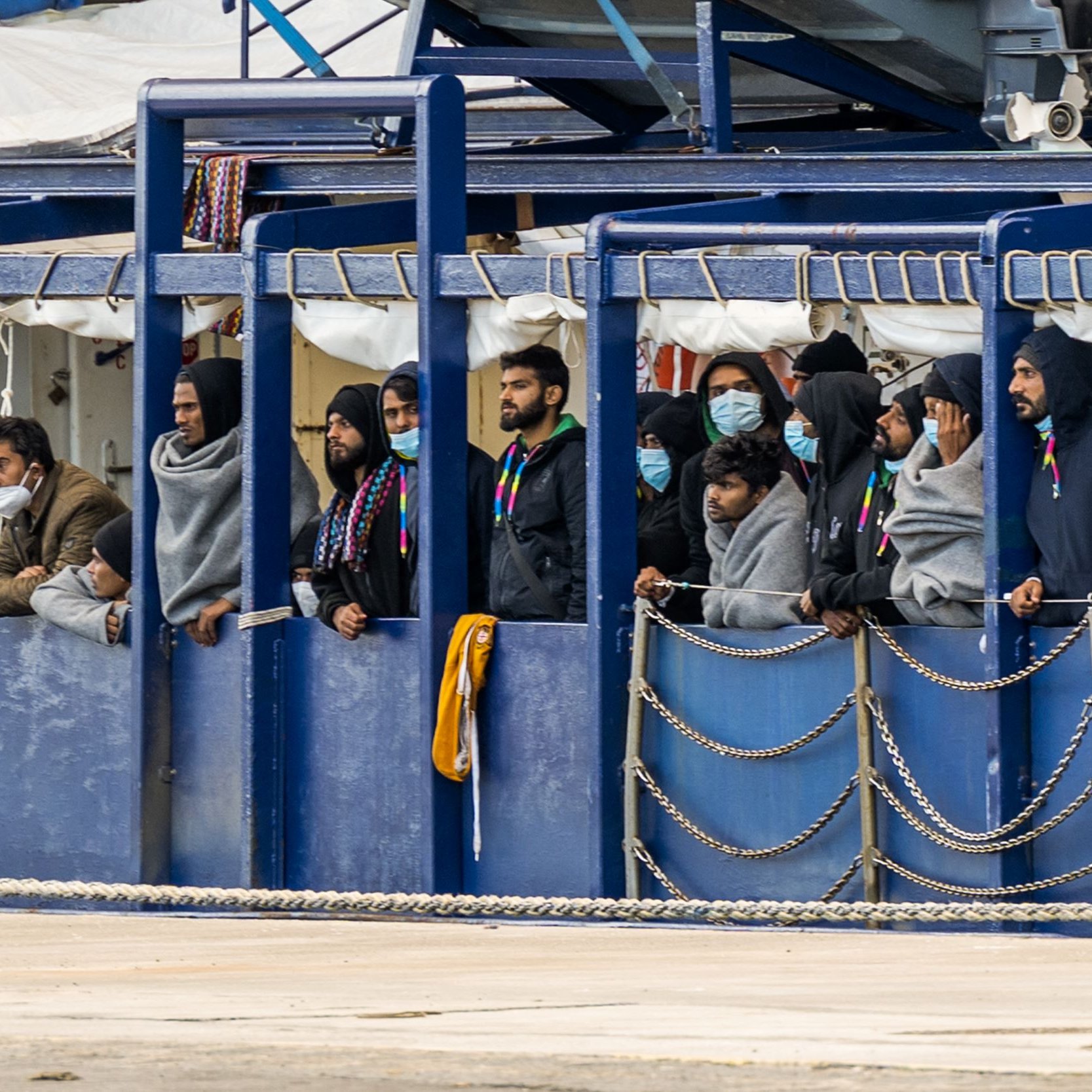 L'image montre un groupe de personnes se tenant sur un bateau. Elles portent des masques et des vêtements semblant chauds, comme des sweats à capuche. Les gens regardent vers l'extérieur, probablement vers la terre ou vers un port. L'environnement semble portuaire, avec des équipements et d'autres structures en arrière-plan. L'atmosphère peut sembler préoccupante ou attendue, reflétant une situation particulière.