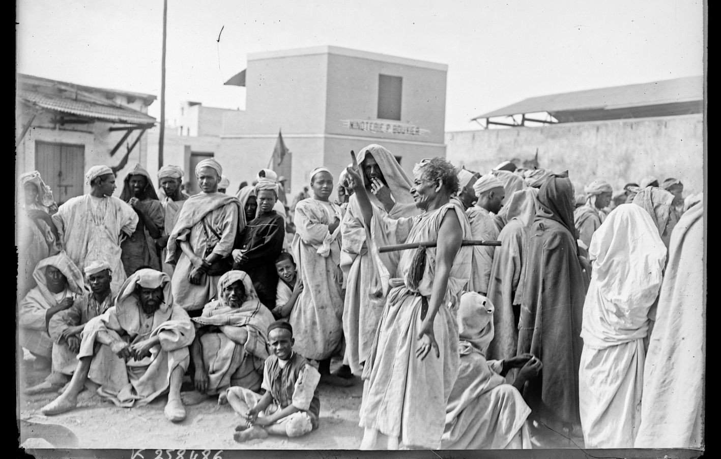 La imagen muestra a un grupo de personas en un entorno que parece ser un mercado o una reunión al aire libre. La mayoría de las personas están vestidas con ropas tradicionales, muchas de las cuales son de colores claros y están cubiertas con prendas que podrían indicar el clima cálido de la región. Hay un enfoque particular en una figura central que parece estar gesticulando o hablando, rodeada de un público atento. El ambiente es de interacción social y hay una variedad de expresiones en los rostros de las personas, que van desde la curiosidad hasta la concentración. El fondo muestra edificaciones simples, lo que sugiere un contexto urbano o rural.
