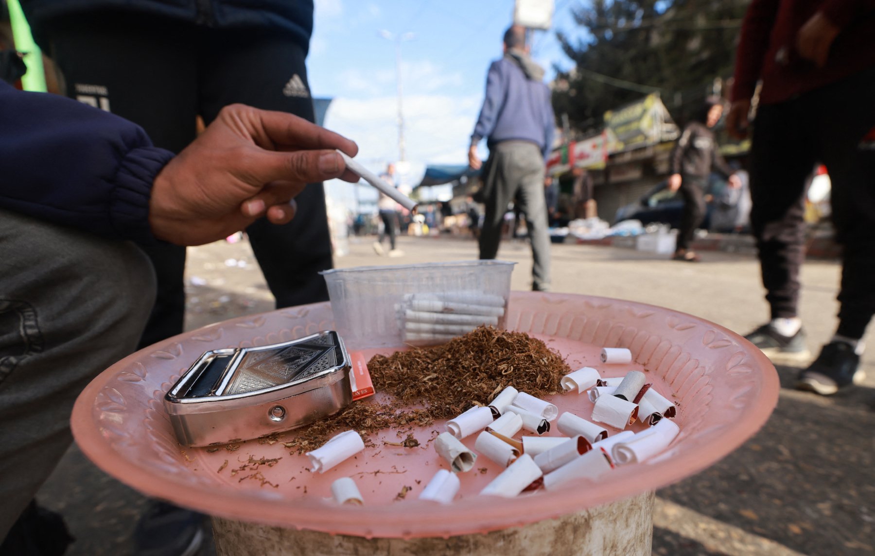 L'immagine mostra una persona che tiene in mano una sigaretta su un piatto di plastica rosa, sul quale sono visibili numerosi mozziconi di sigaretta. Accanto ai mozziconi c'è del tabacco sfuso e un contenitore trasparente. Lo sfondo rivela una strada affollata, con persone che si muovono e diversi cartelloni pubblicitari. L'atmosfera sembra quella di un mercato o di una zona urbana.