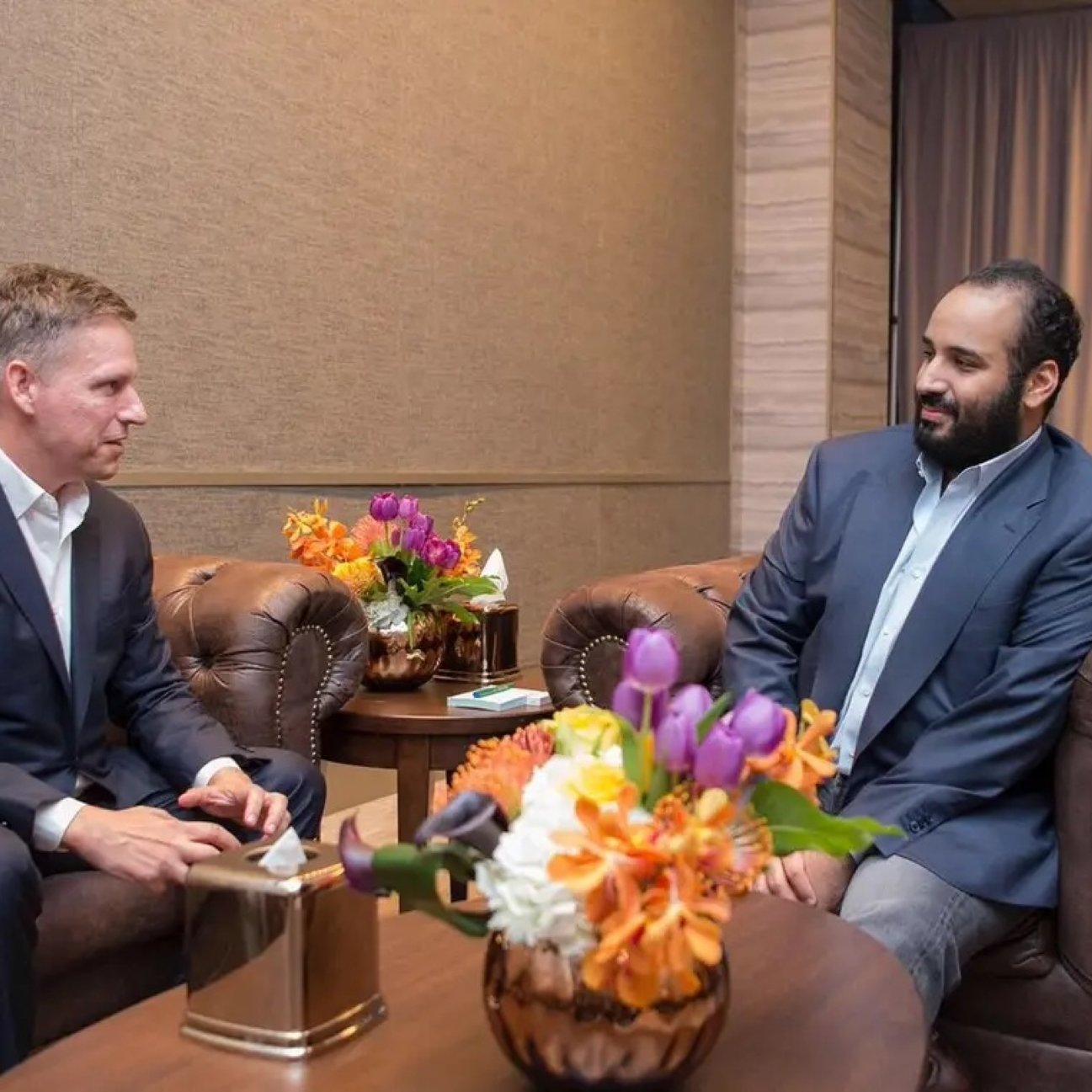 Deux hommes en costume assis dans un salon, entourés de fleurs colorées.