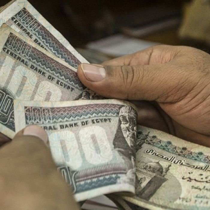 L'image montre des mains tenant des billets égyptiens. On peut voir des coupures de différentes valeurs, principalement de 100 livres égyptiennes. Les billets sont colorés et comportent des motifs et des inscriptions en arabe. L'arrière-plan semble flou, indiquant que l'attention est centrée sur l'argent.