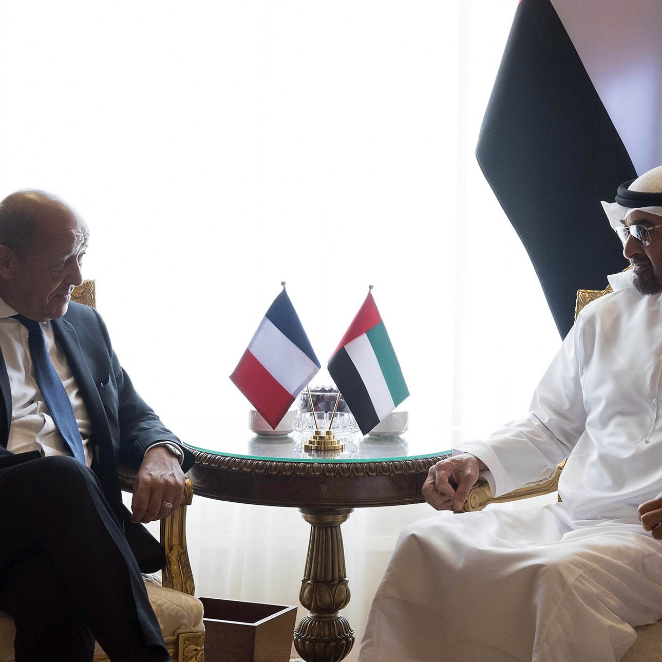 L'image montre deux hommes assis sur des fauteuils dans un cadre élégant. Ils discutent autour d'une petite table portant des drapeaux de la France et des Émirats Arabes Unis. L'un des hommes est vêtu d'un costume, tandis que l'autre porte une tenue traditionnelle blanche. Leurs expressions semblent amicales et engageantes, suggérant une conversation diplomatique. L'arrière-plan est décoré de rideaux et d'autres éléments luxueux.