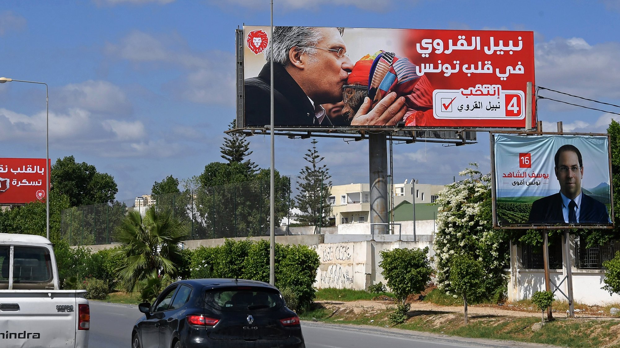 Sur cette image, on voit un panneau publicitaire en arabe. Le panneau montre deux personnages qui s'embrassent, entourés de couleurs vives et de slogans. En bas, on peut voir une photo d'une autre personne (peut-être un candidat) avec un texte supplémentaire à son propos. La scène se déroule dans un environnement urbain, avec des voitures circulant sur une route. Le ciel est bleu, parsemé de nuages.