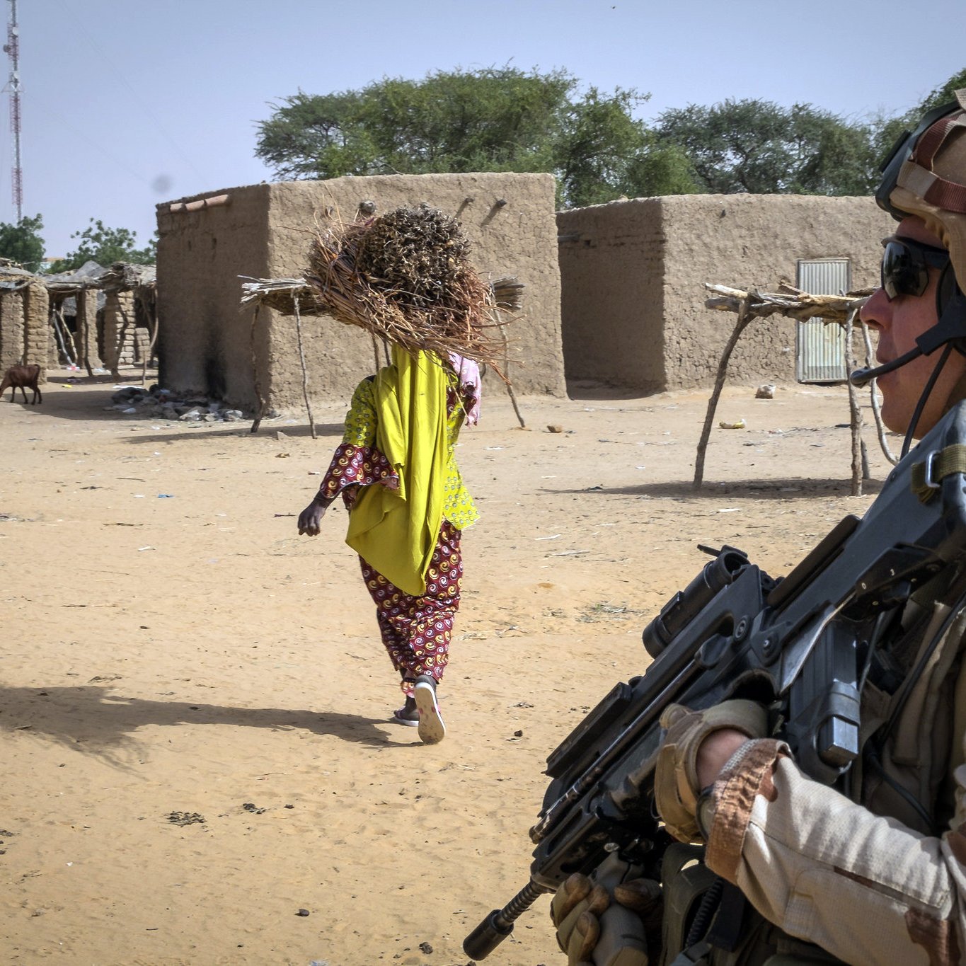 The image depicts a soldier standing in the foreground, holding a rifle and wearing military gear, including a helmet and camouflage uniform. The soldier appears vigilant as they face away from the camera. In the background, there is a woman walking away, carrying a large bundle of sticks on her head, dressed in a brightly colored traditional outfit. The setting looks like a rural area with simple mud-brick structures and some sparse vegetation, indicating a dry environment. The overall scene suggests a blend of military presence and civilian life in a remote location.