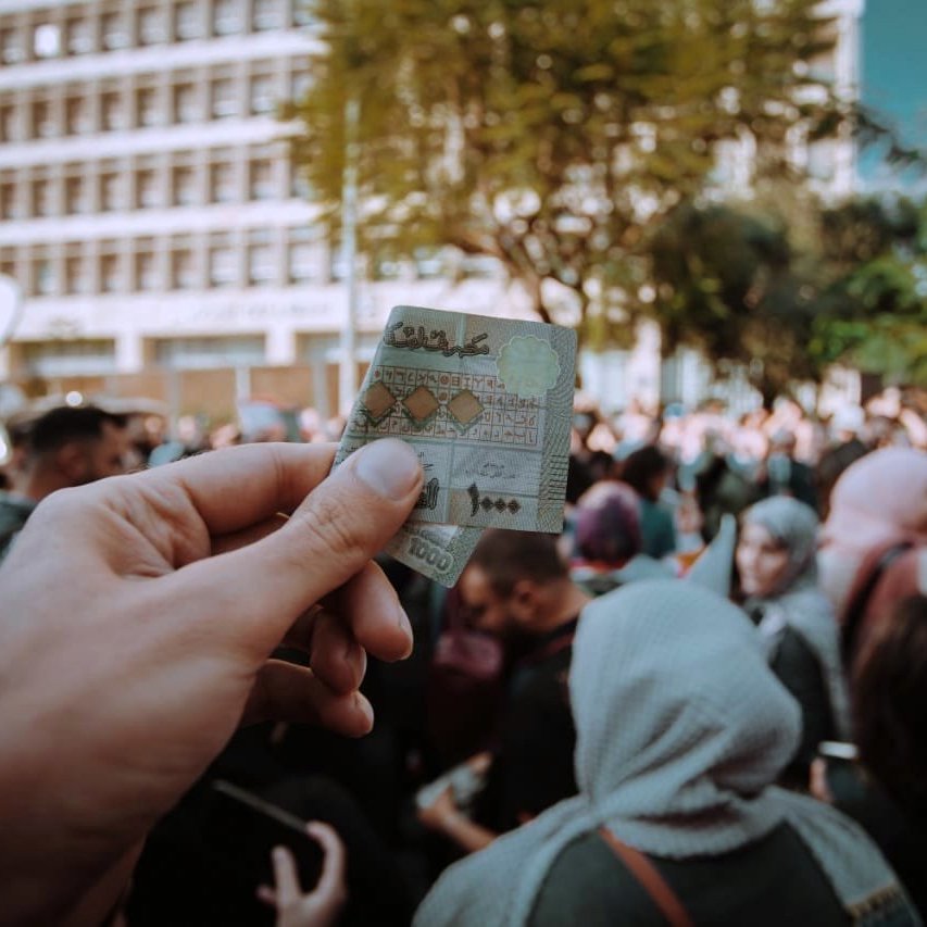 L'image montre une main tenant un billet de banque marocain au premier plan, tandis qu'à l'arrière-plan, une foule est rassemblée. On aperçoit des personnes aux tenues variées, certaines portant des foulards. Le cadre semble être un événement public, probablement une manifestation ou une célébration, avec des bâtiments modernes en arrière-plan et des arbres qui ajoutent une touche de verdure à la scène. L'atmosphère semble dynamique et chargée d'énergie collective.