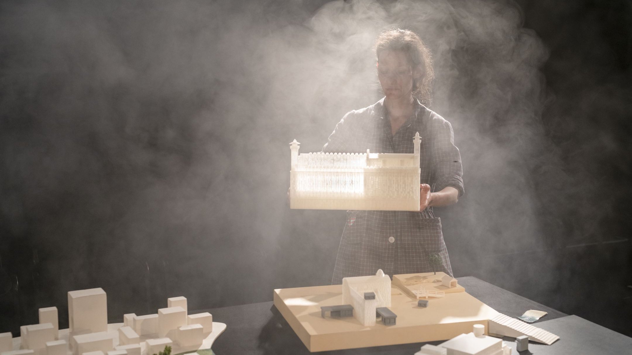 L'image montre une personne tenant une maquette d'un bâtiment, dans un environnement brumeux. Le fond est sombre, ce qui met en valeur la maquette et la silhouette de la personne. Sur la table, il y a d'autres maquettes et éléments architecturaux, suggérant une scène de travail ou de présentation liée à l'architecture. La lumière semble filtrer à travers la brume, créant une ambiance mystérieuse et artistique.