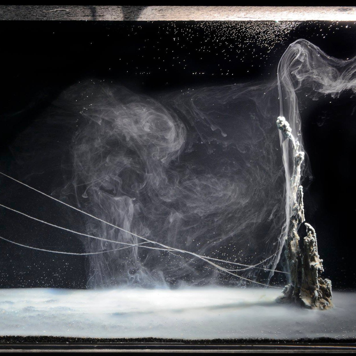 The image showcases a mesmerizing underwater scene featuring swirling currents and bubbles. The foreground has a smooth, almost glass-like surface with a hint of foam, while the background features abstract formations that resemble smoke or flowing water. Thin, delicate tendrils or strings extend across the scene, creating an ethereal atmosphere. The overall color palette is dominated by dark tones, emphasizing the luminous qualities of the water and creating a sense of depth and motion.