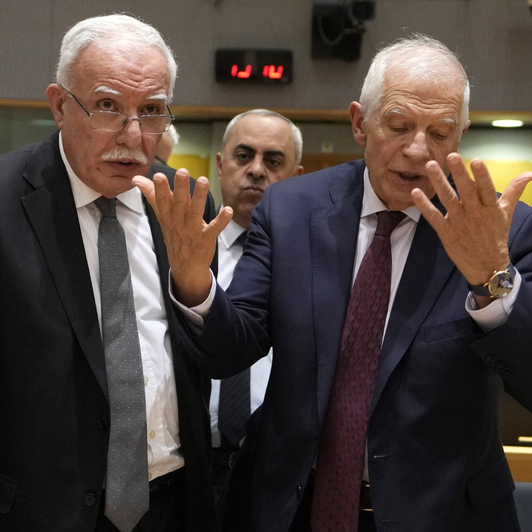 L'image montre deux hommes en discussion animée, debout dans un cadre de réunion. L'un d'eux a des cheveux gris et porte des lunettes, tandis que l'autre est également âgé avec des cheveux clairs. Ils semblent gesticuler avec leurs mains pour souligner leurs propos. En arrière-plan, une autre personne les observe. L'environnement paraît être une salle de conférence, avec des tables et des collègues travaillant.