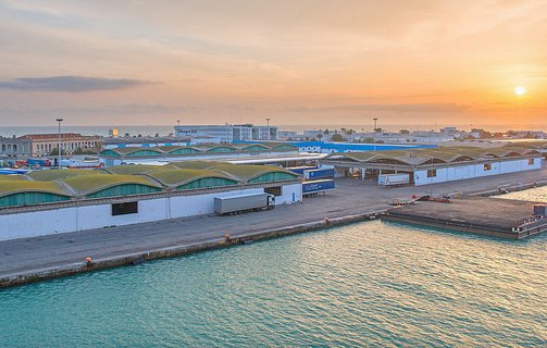 L'image montre un port lors d'un coucher de soleil, avec un paysage industriel. On peut voir des entrepôts aux toits arrondis, bordés par l'eau. Des véhicules et des conteneurs sont visibles, indiquant une activité portuaire. Le ciel est teinté de couleurs chaudes, créant une atmosphère paisible et accueillante.