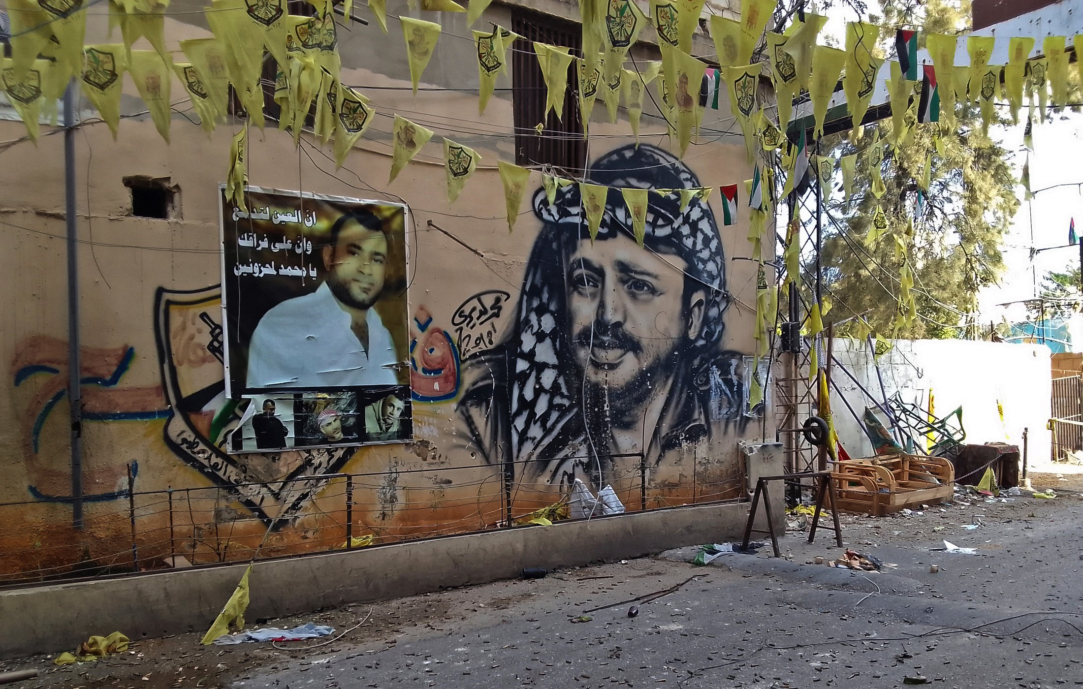 L'image montre un mur décoré d'une grande fresque représentant un homme avec un keffieh, typique de la culture arabe. À côté, il y a des affiches avec des portraits et des slogans. Des drapeaux jaunes pendent également, créant une ambiance festive ou militante. Le sol semble être en désordre avec divers objets autour, indiquant peut-être un événement ou une célébration récente. Les couleurs vives des décorations contrastent avec le mur beige.