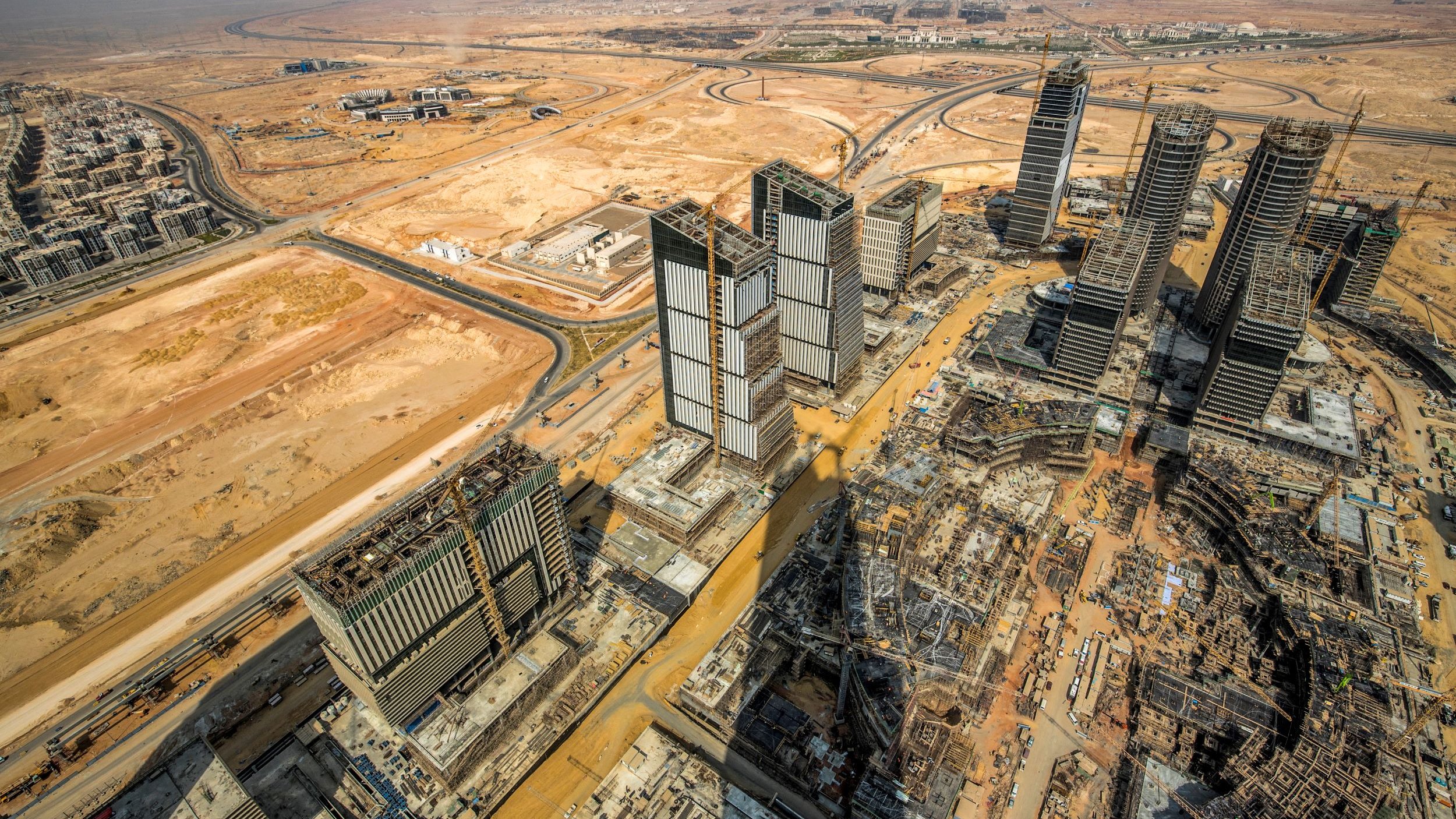 The image showcases a panoramic view of a vast construction site in a desert landscape. In the foreground, several high-rise buildings are partially completed, with cranes and construction equipment evident. The surrounding area features empty land and some roadways, characteristic of a developing urban environment. The backdrop includes a clear sky and distant structures, suggesting a mixture of ongoing urbanization and natural desert terrain. The overall scene illustrates the rapid expansion of urban infrastructure in a typically arid region.