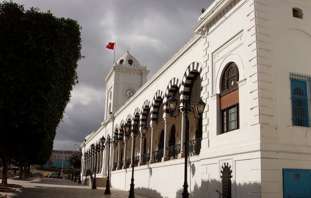 تظهر الصورة مبنىً تاريخياً يتميز بالتصميم المعماري الأنيق. يتمتع المبنى بجدران بيضاء وأسقف مميزة، ويحتوي على نوافذ كبيرة مزينة بأقواس. هناك ساعة كبيرة على أحد جوانب المبنى، وعلوه يبرز به علم تركيا. تحيط بالمكان أشجار خضراء، بالإضافة إلى ممرات مبلطة وأعمدة إنارة تضفي جواً جمالياً على المشهد. السماء تظهر محملة بالغيوم، مما يضيف طابعاً درامياً إلى الصورة.