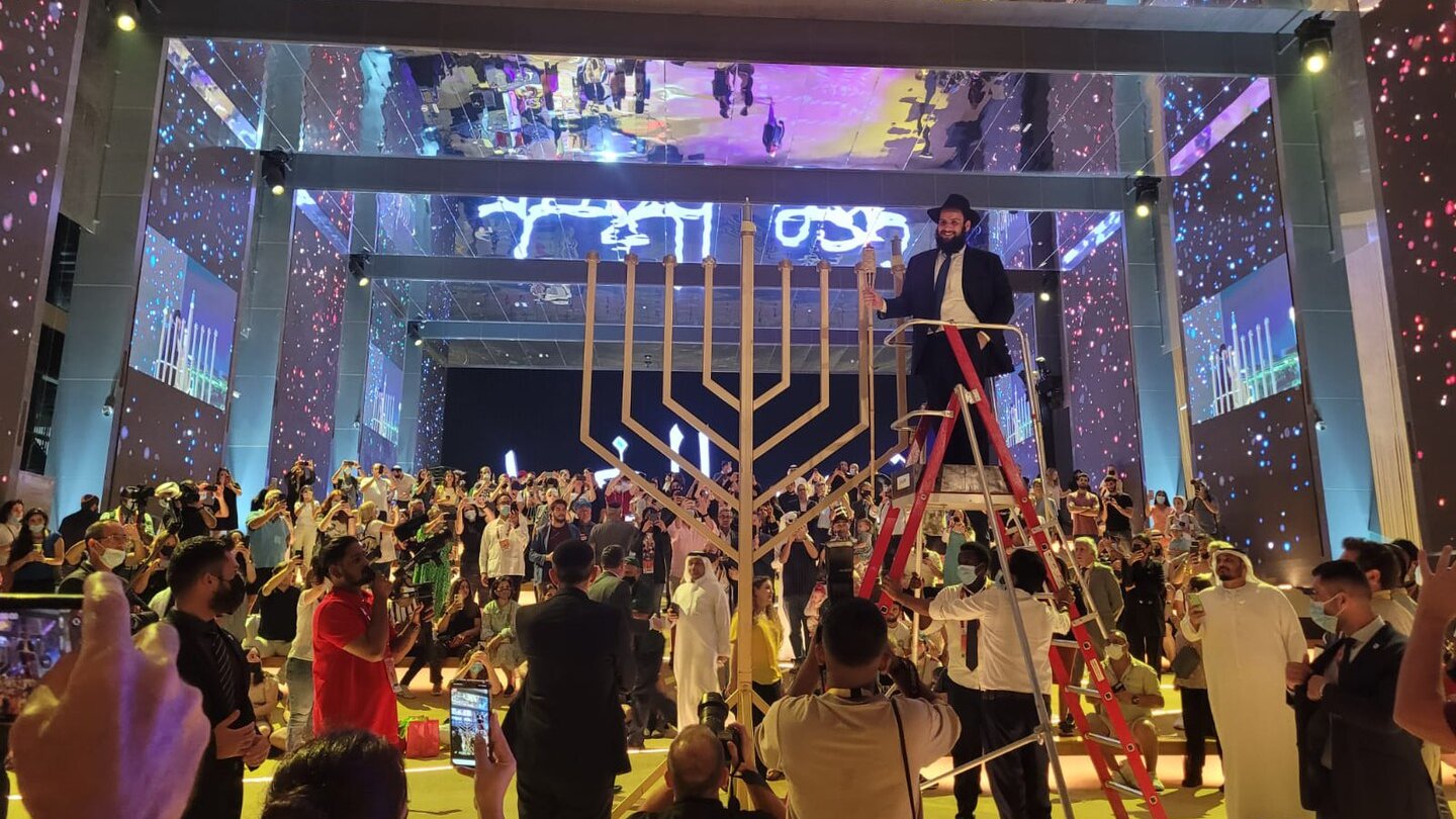 The image depicts a lively event taking place in a large, well-lit venue. In the center, there is a large menorah, which is a symbol associated with Hanukkah. A person is standing on a ladder, seemingly preparing to light the menorah. Surrounding the menorah, a diverse crowd is gathered, with some individuals clapping or taking photos. The background features colorful lights and projections, enhancing the festive atmosphere of the gathering. The overall scene reflects a sense of celebration and community.