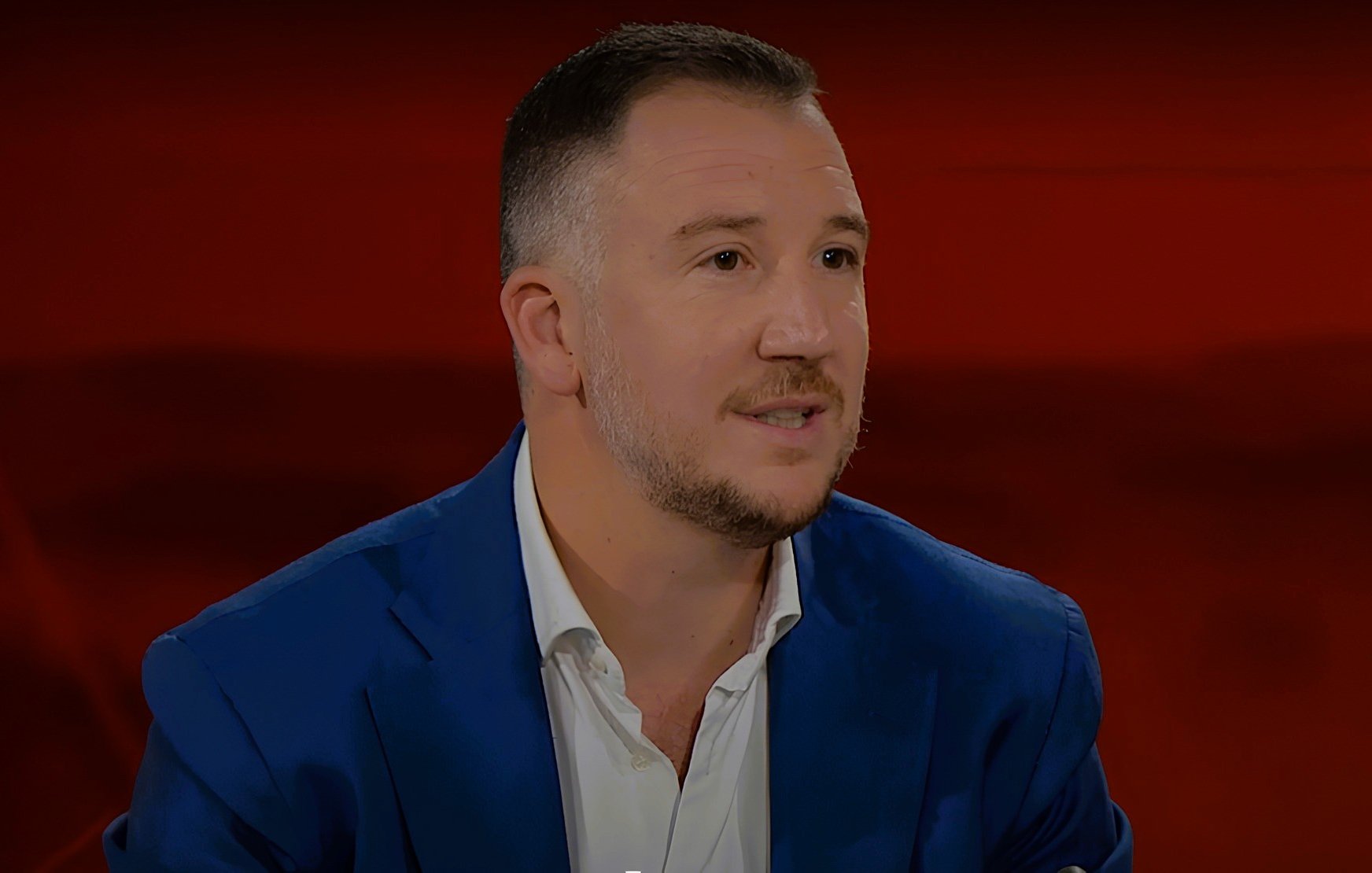 L'immagine mostra un uomo con capelli corti e una barba leggera, che indossa una giacca blu e una camicia bianca. Si trova di fronte a uno sfondo rosso. L'uomo appare in un contesto di colloquio o discussione, con un'espressione attenta e coinvolta.