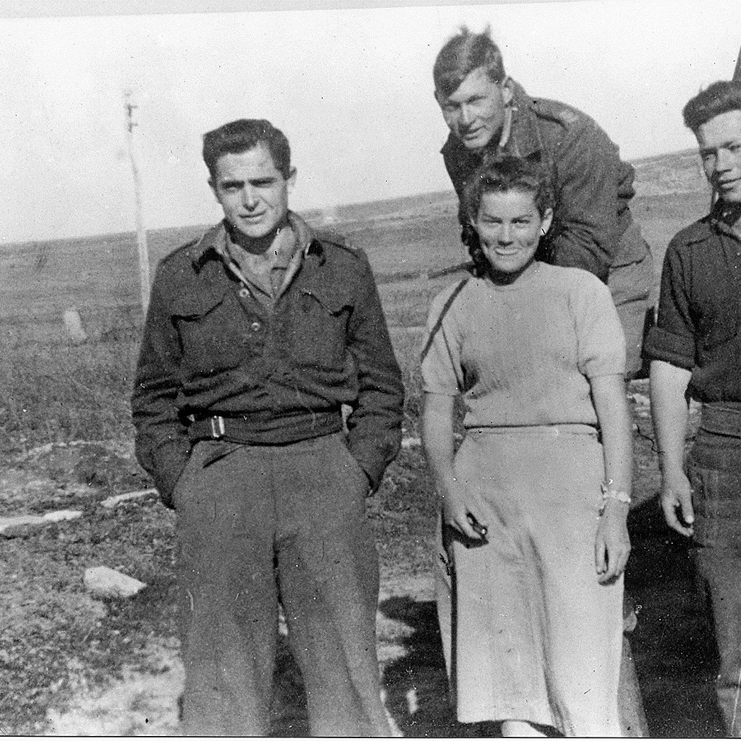 L'image montre quatre personnes se tenant debout à l'extérieur, probablement sur un terrain dégagé. Il y a trois hommes et une femme. Les hommes portent des uniformes militaires ou des vêtements utilitaires, tandis que la femme est habillée simplement dans une robe. L'environnement semble rural, avec une petite bâtisse en arrière-plan et des champs en toile de fond. Tout le monde affiche des expressions neutres ou souriantes, créant une atmosphère de camaraderie.