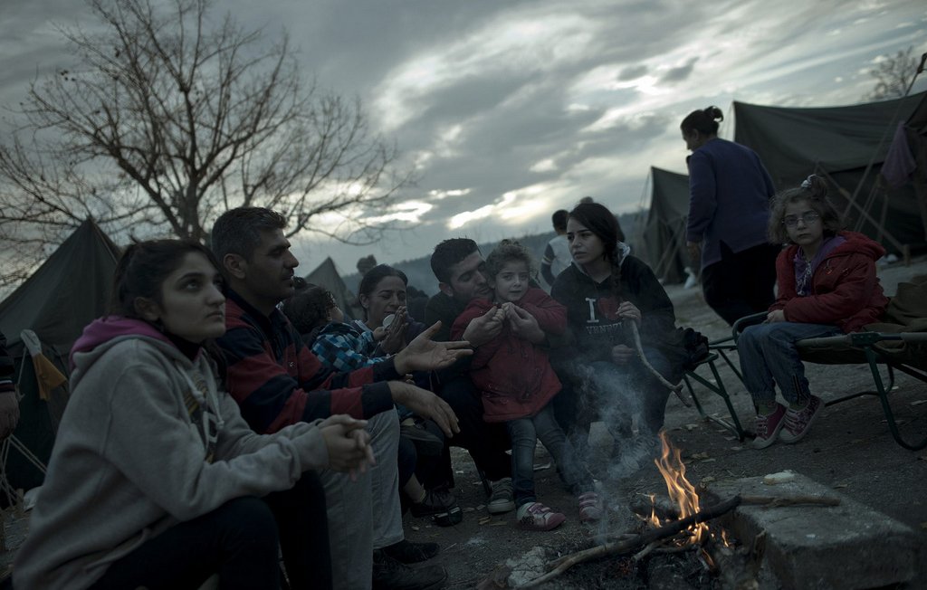 L'image montre un groupe de personnes assises autour d'un feu de camp, probablement dans un camp de réfugiés ou un site de fortune. On peut voir des adultes et des enfants, certains vêtus de manteaux pour se protéger du froid. L'ambiance semble sombre en raison des nuages menaçants dans le ciel. Les visages des personnes expriment une variété d'émotions, allant de la tristesse à l'espoir. En arrière-plan, on aperçoit des tentes qui indiquent que ce groupe vit dans des conditions temporaires.