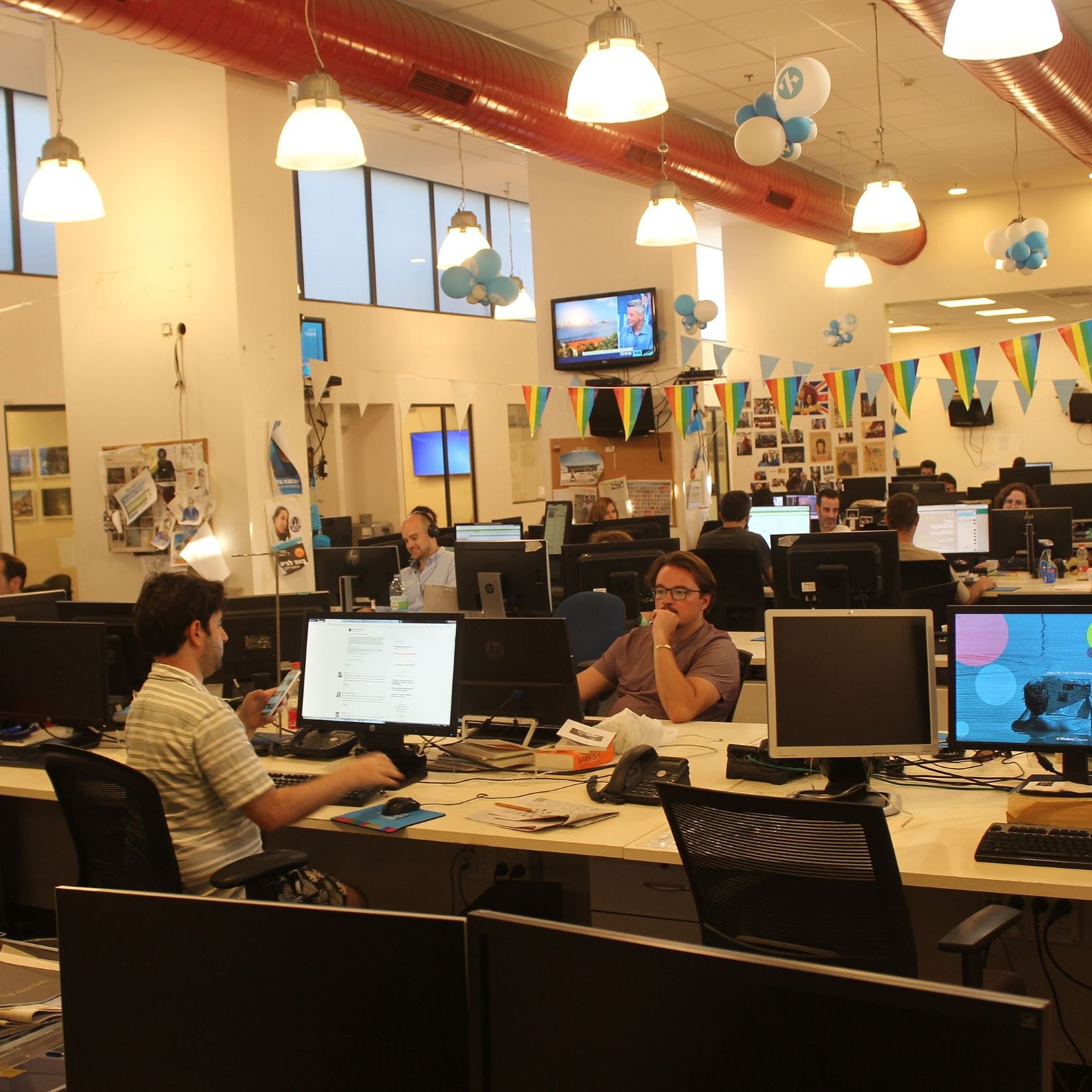 L'image montre un environnement de bureau moderne et animé. Plusieurs employés sont assis devant des ordinateurs, travaillant sur leurs tâches. Les bureaux sont équipés de plusieurs écrans d'ordinateur. La pièce est décorée avec des ballons et des banderoles, suggérant une atmosphère festive ou un événement spécial. La lumière naturelle entre par de grandes fenêtres, ajoutant une ambiance agréable à l'espace de travail.