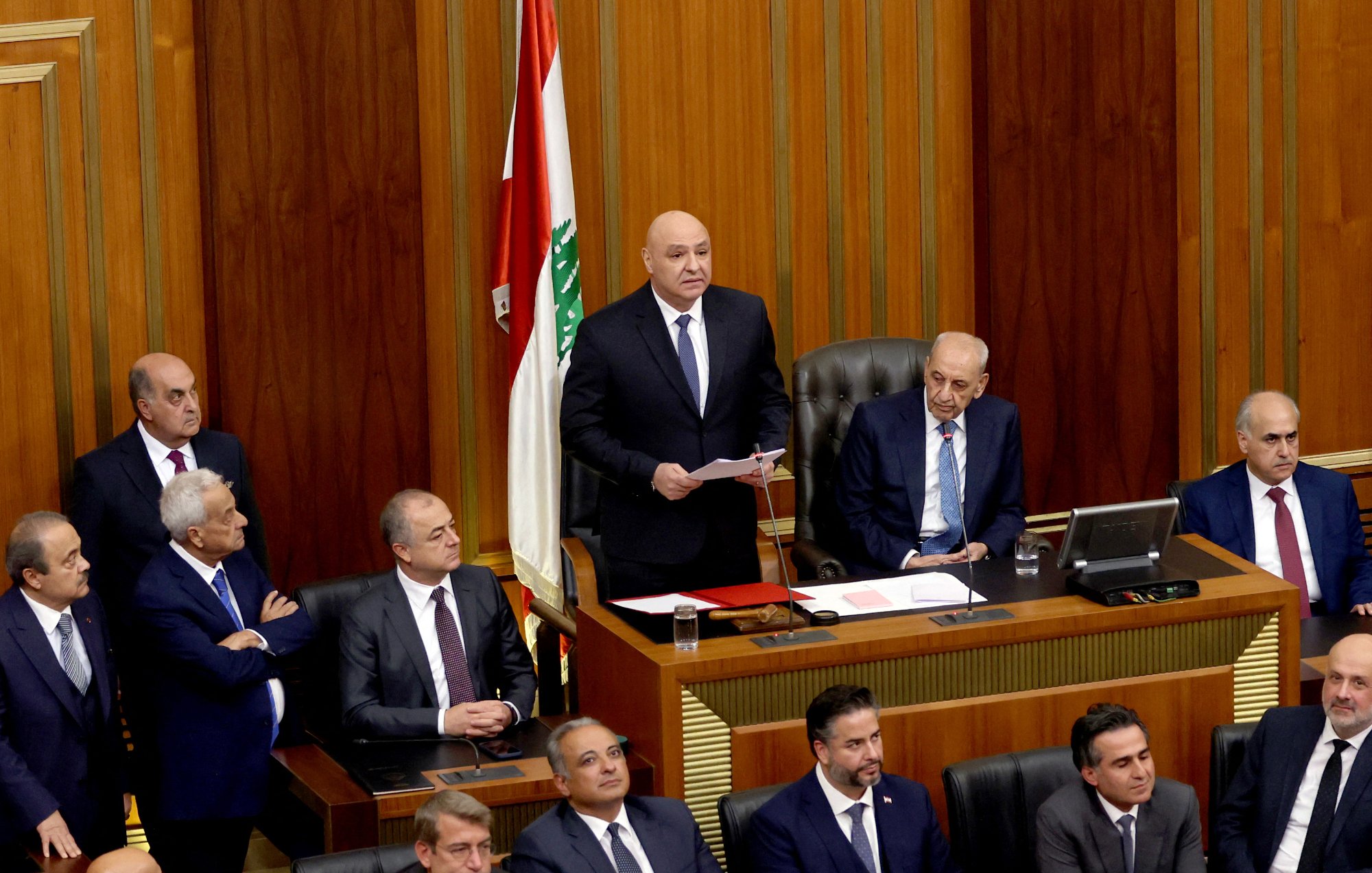 این تصویر یک جلسه رسمی در مجلس لبنان را نشان می‌دهد. در مرکز تصویر، یک فرد در حال سخنرانی است و تعدادی دیگر از اعضای مجلس در اطراف او نشسته‌اند. رومیزی مجلس با مستنداتی پوشیده شده و یک پرچم لبنان نیز در پس‌زمینه قابل مشاهده است. به نظر می‌رسد که جو جلسه جدی و رسمی است.