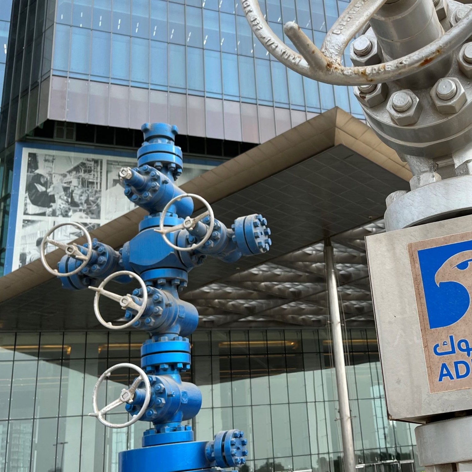 L'image montre un bâtiment moderne avec une façade en verre. À l'avant, on distingue deux équipements de contrôle, généralement utilisés dans le secteur pétrolier. L'un des équipements est de couleur bleue, tandis que l'autre est gris et porte un logo au sommet indiquant "ADNOC", qui est l'acronyme de la Compagnie Pétrolière Nationale des Émirats Arabes Unis. On peut également voir des panneaux avec le nom ADNOC en arabe et en anglais.