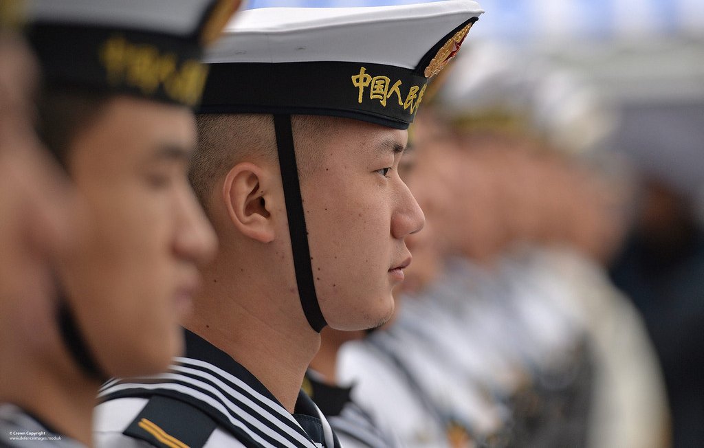 L'image montre des marins en uniforme, probablement lors d'une cérémonie ou d'un événement officiel. On peut voir des hommes alignés, portant des chapeaux distinctifs et des uniformes marins. Les expressions sur leurs visages sont sérieuses, et ils semblent concentrés sur un point donné. L'arrière-plan montre des silhouettes floues d'autres marins, suggérant un grand rassemblement. L'image dégage une ambiance de discipline et de camaraderie militaire.