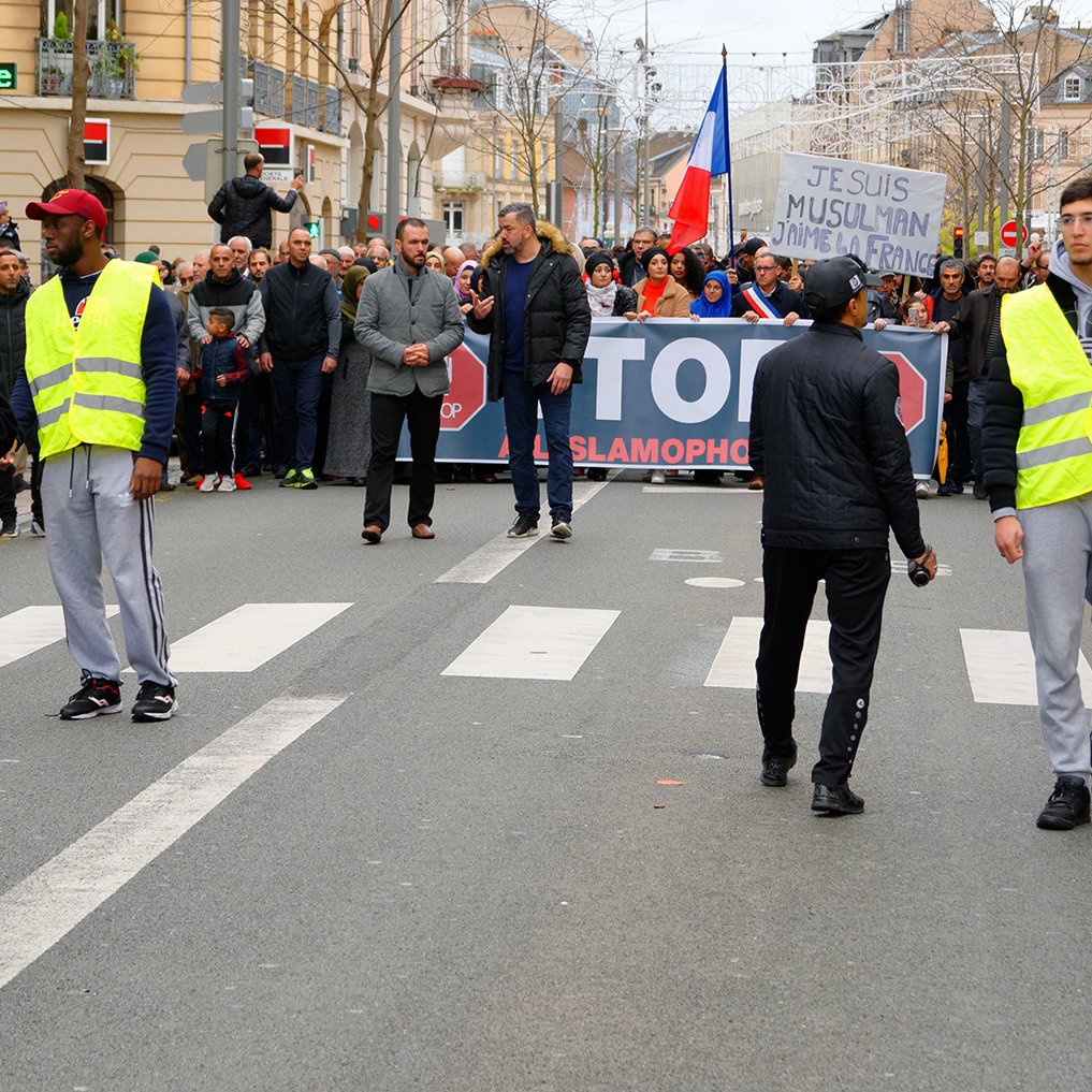 L'image montre une manifestation dans une rue où un groupe de personnes tient une grande banderole avec des slogans. Au premier plan, deux agents de sécurité ou policiers portent des gilets réfléchissants et semblent surveiller la situation. Des manifestants sont visibles à l'arrière, et il y a une ambiance de mobilisation avec des drapeaux. L'environnement urbain se présente avec des bâtiments voisins et une circulation routière réduite à ce moment.