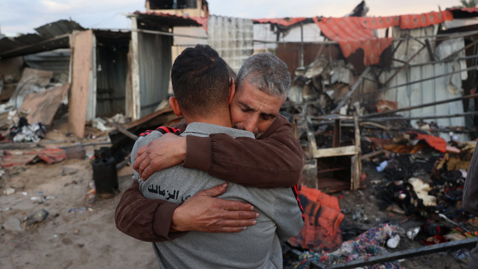 L'image montre deux hommes qui s'étreignent de manière émotive au milieu de décombres. En arrière-plan, il y a des restes de bâtiments détruits, avec des morceaux de métal et des débris éparpillés. L'ambiance semble très chargée d'émotion, suggérant une perte ou un chagrin. Les expressions des hommes témoignent d'une connexion forte et d'un besoin de réconfort dans un moment difficile.