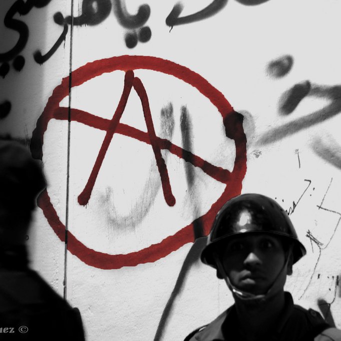 L'image présente un mur recouvert de graffitis, dont un symbole anarchiste stylisé en rouge, entouré d'un cercle. En arrière-plan, on voit deux hommes en uniforme, probablement des forces de l'ordre. L'un d'eux est au premier plan, portant un casque, avec une expression sérieuse. L'atmosphère de la photo suggère une tension sociale, accentuée par l'utilisation de couleurs sombres et le contraste avec le symbole rouge.