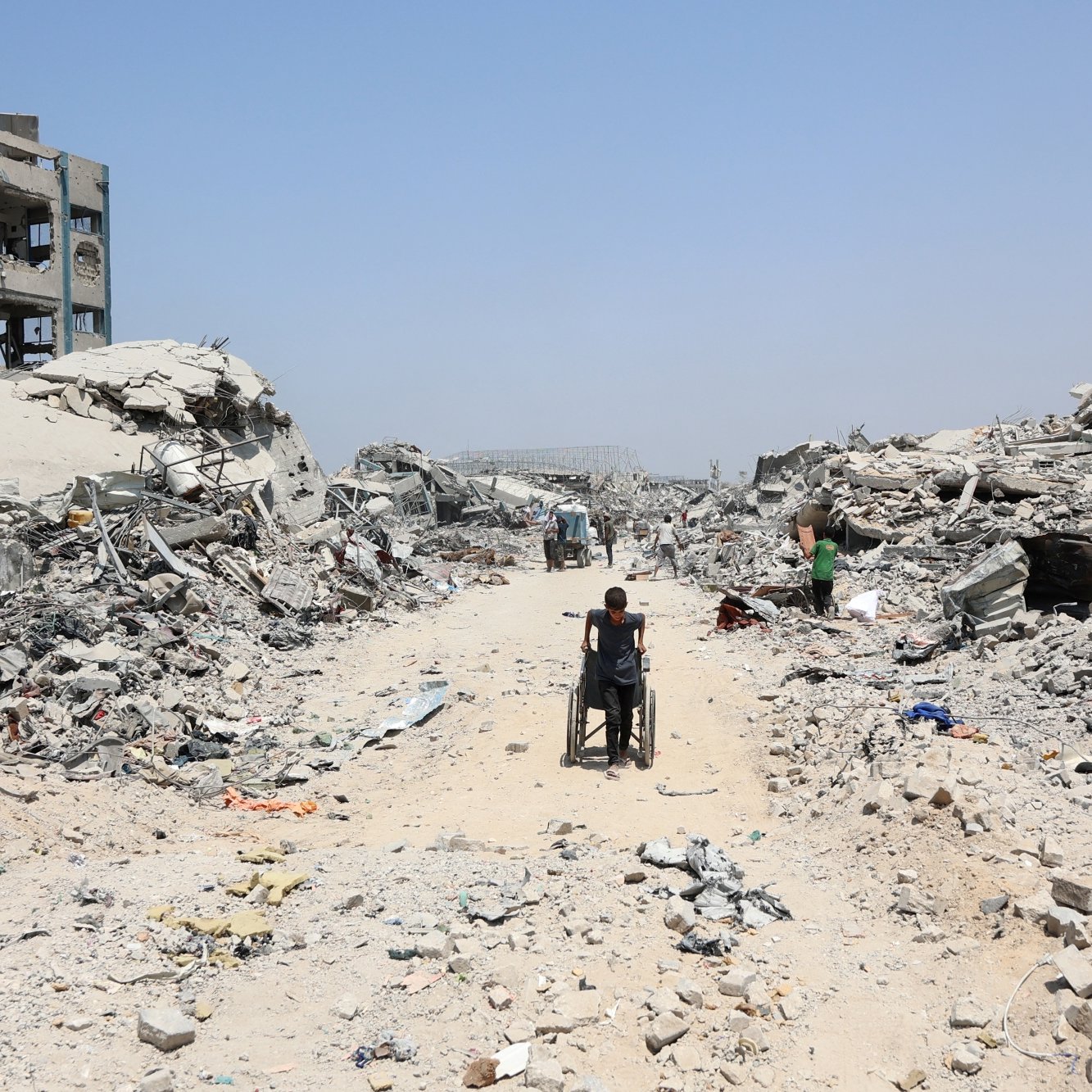 Una strada polverosa tra macerie di edifici distrutti, con una persona in sedia a rotelle.