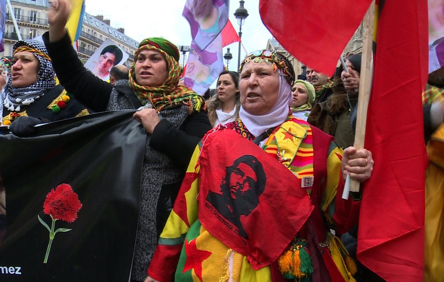 L'image montre une manifestation avec des personnes brandissant des drapeaux et des bannières. Certaines femmes portent des vêtements traditionnels et des accessoires colorés, et elles expriment leurs revendications avec des gestuelles passionnées. Il y a également des portraits visibles en arrière-plan, suggérant un hommage à des individus particuliers. L'ambiance semble être à la fois festive et engagée, avec une forte symbolique culturelle.