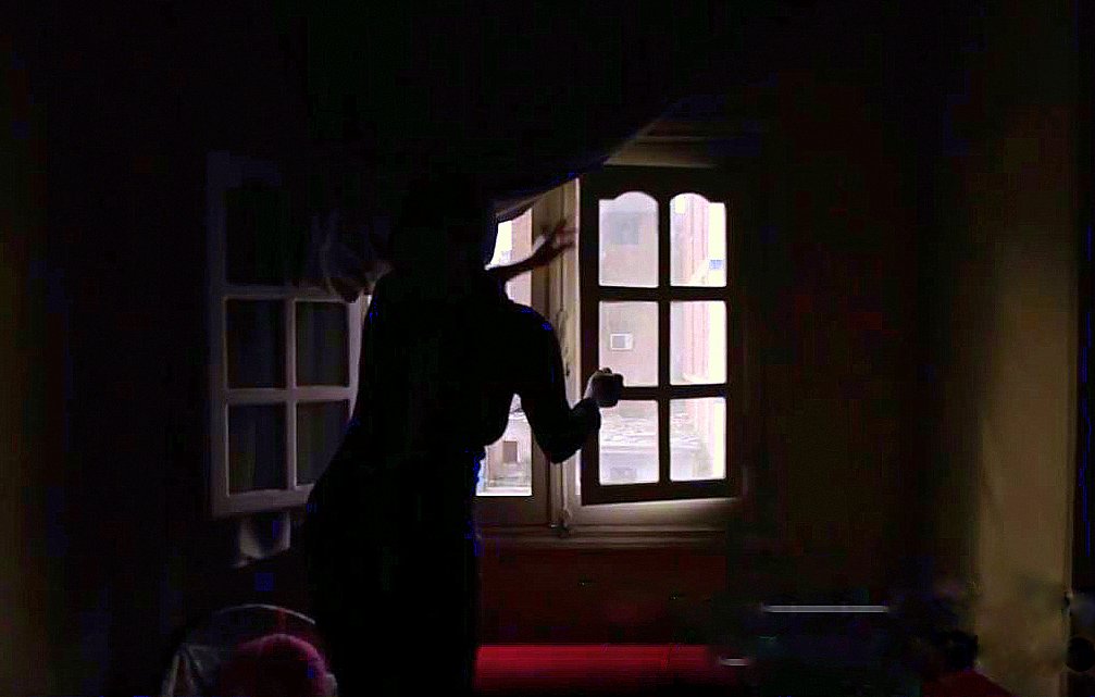 L'image montre une silhouette se tenant près d'une fenêtre dans une pièce sombre. La lumière entre par la fenêtre, illuminant partiellement l'espace environnant, tandis que des détails tels que les murs et les meubles sont à peine visibles dans l'ombre. La silhouette semble en mouvement, ajoutant une ambiance dynamique et mystérieuse à la scène. Les rideaux sont légèrement ouverts, laissant entrevoir l'extérieur.