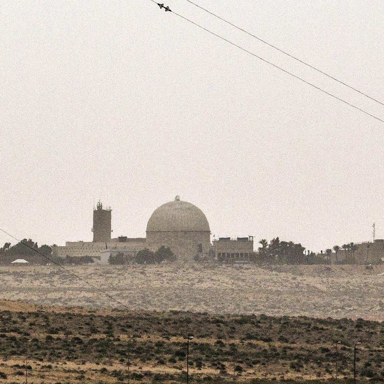 L'image montre un paysage désertique avec un complexe architectural. On peut voir une grande structure arrondie, probablement un dôme, qui se démarque dans le ciel brumeux. À proximité, il y a une cheminée avec des bandes de couleur, suggérant une activité industrielle. Le terrain environnant est aride, avec quelques arbres dispersés. L'atmosphère semble légèrement brumeuse, ce qui donne une ambiance calme et presque mystérieuse à la scène.