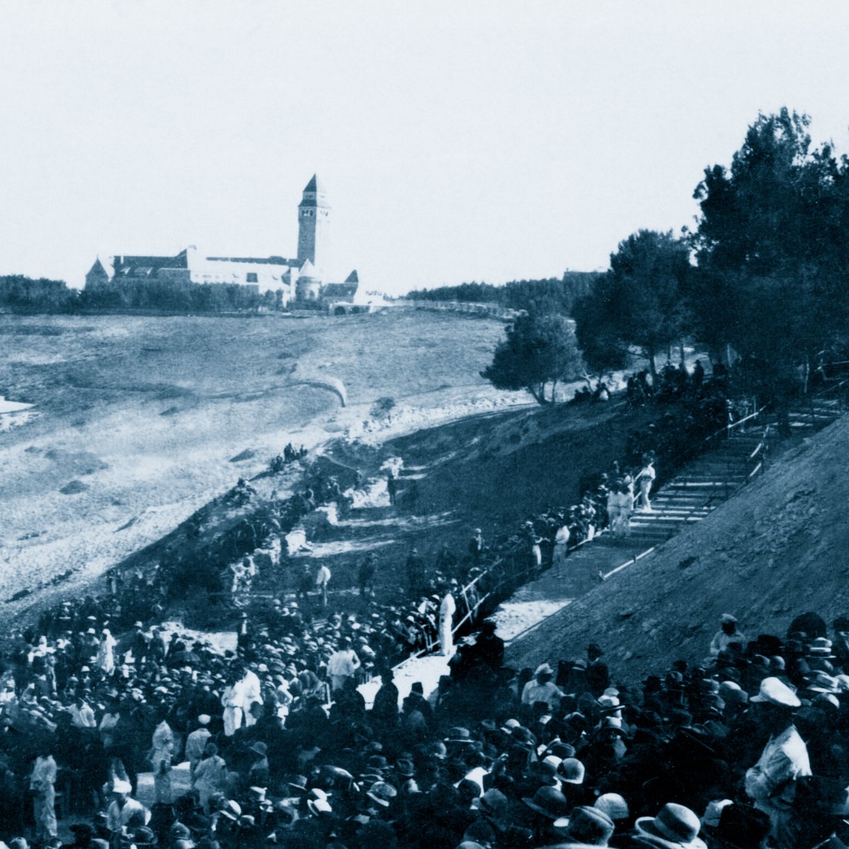 Un grande raduno di persone all'aperto, con un edifico distante e alberi circostanti.