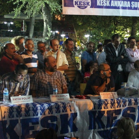 L'image montre une scène de rassemblement ou de meeting en extérieur. Une table est placée au premier plan, où plusieurs personnes sont assises, apparemment des intervenants, pendant qu'une autre personne, debout, s'adresse à l'audience. Un public se tient autour, écoutant attentivement. En arrière-plan, il y a une bannière avec des inscriptions, et l'ambiance semble être celle d'un événement communautaire ou d'une manifestation. Les participants semblent engagés dans une discussion animée.