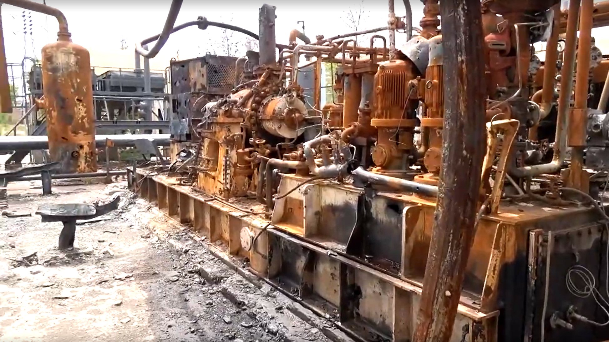 L'image montre une installation industrielle endommagée, probablement suite à un incendie. On peut voir des équipements métalliques, tels que des machines et des tuyaux, recouverts de rouille et de suie. Le sol est noirci par des résidus, indiquant les conséquences d'un sinistre. L'environnement semble très dégradé, avec des éléments de construction en arrière-plan également affectés par des dommages. L'ensemble donne une impression de désolation et de négligence.