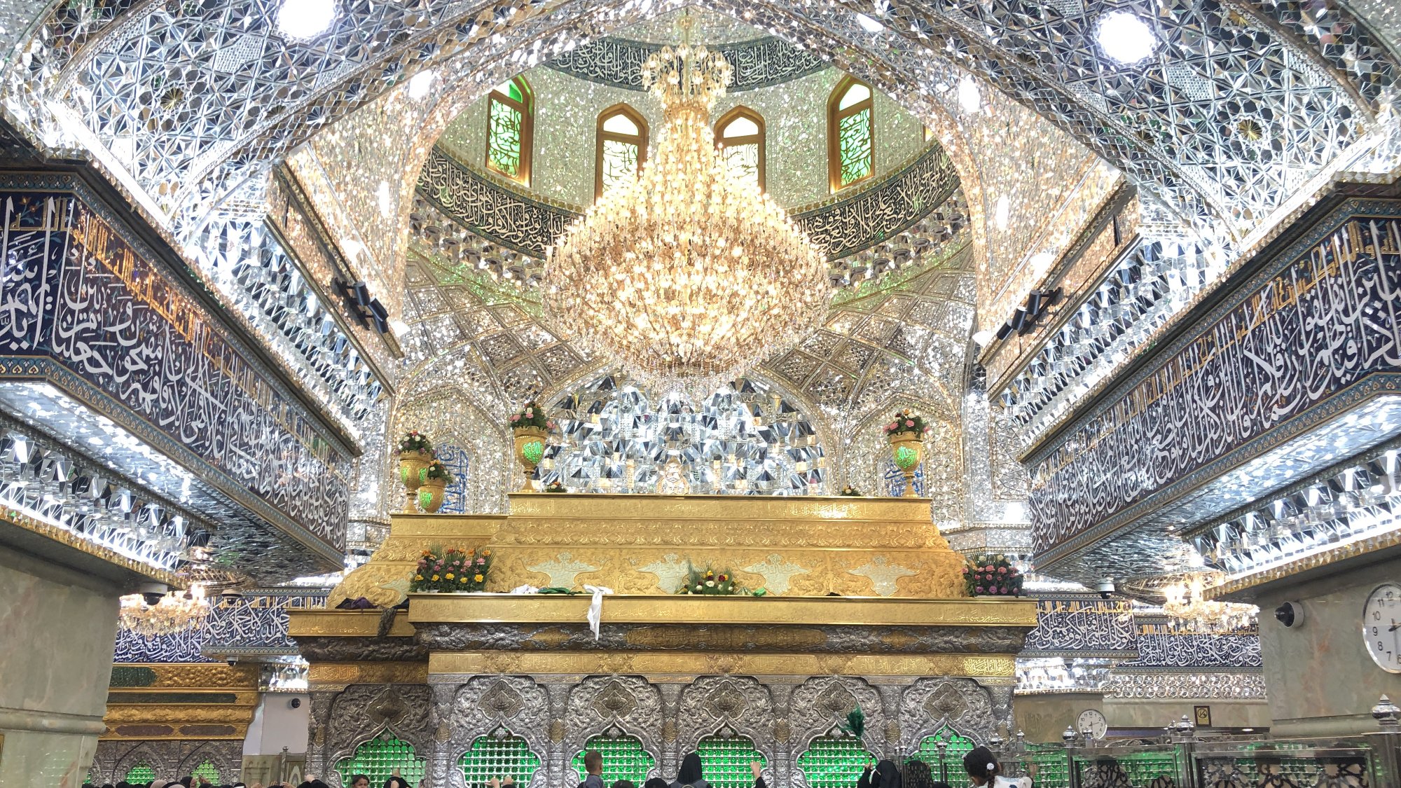 L'image montre un intérieur somptueux, probablement d'un sanctuaire ou d'une mosquée, orné de nombreux détails décoratifs en miroir et de magnifiques motifs. Au centre, on peut voir une structure dorée qui attire l'attention, probablement un mausolée ou une tombe. Le plafond est impressionnant, avec un grand lustre et des fenêtres laissant passer la lumière. Beaucoup de personnes, principalement vêtues de noir, sont rassemblées à l'intérieur, créant une atmosphère de recueillement et de dévotion. Les murs sont également ornés d'inscriptions arabes, ajoutant à la beauté spirituelle du lieu.