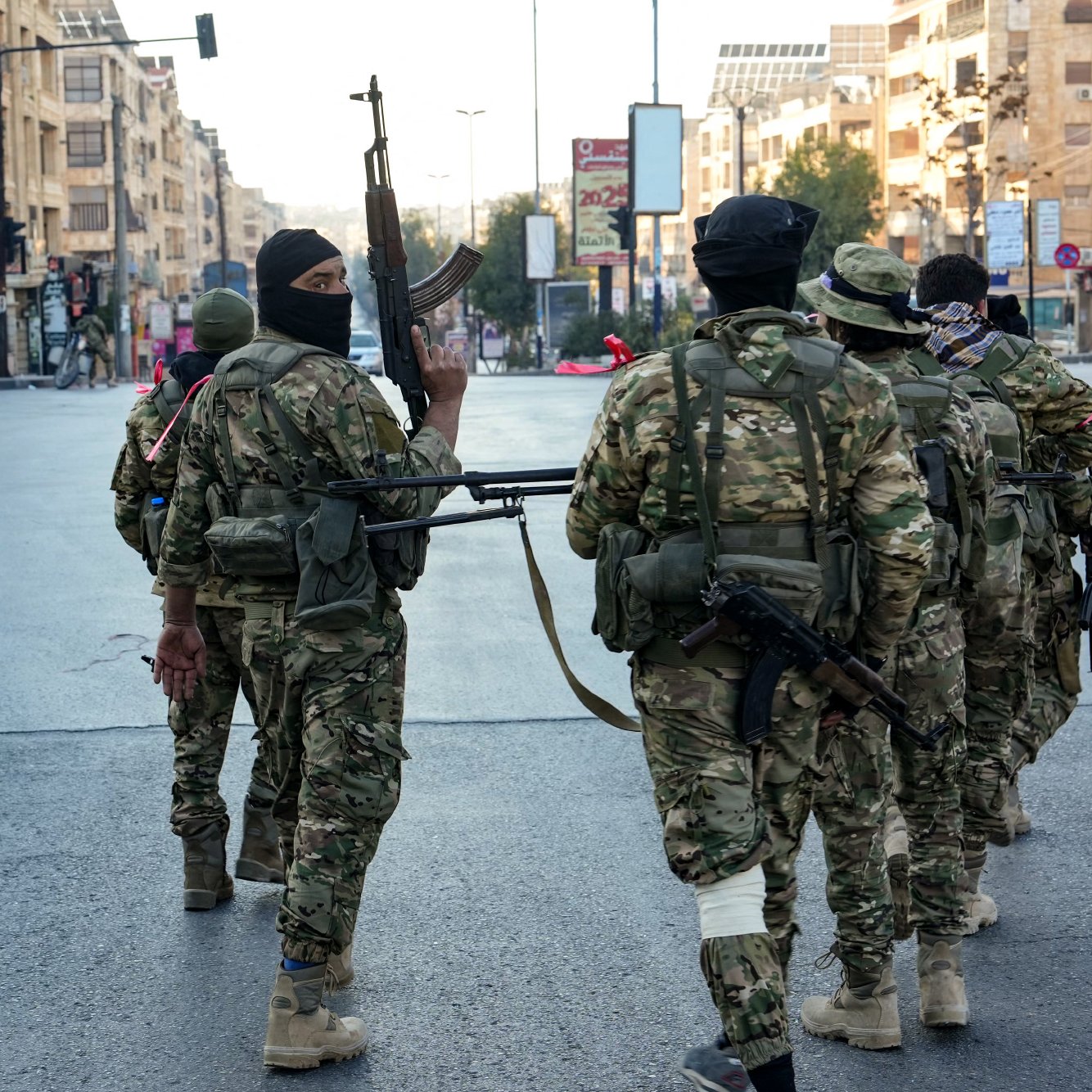 L'image montre un groupe de soldats armés marchant dans une rue urbaine. Ils portent des uniformes militaires et sont équipés d'armes. L'environnement semble urbain, avec des bâtiments en arrière-plan. L'ambiance est sérieuse, suggérant une situation potentiellement tendue. Les soldats portent également des masques, ce qui peut indiquer des préoccupations de sécurité.