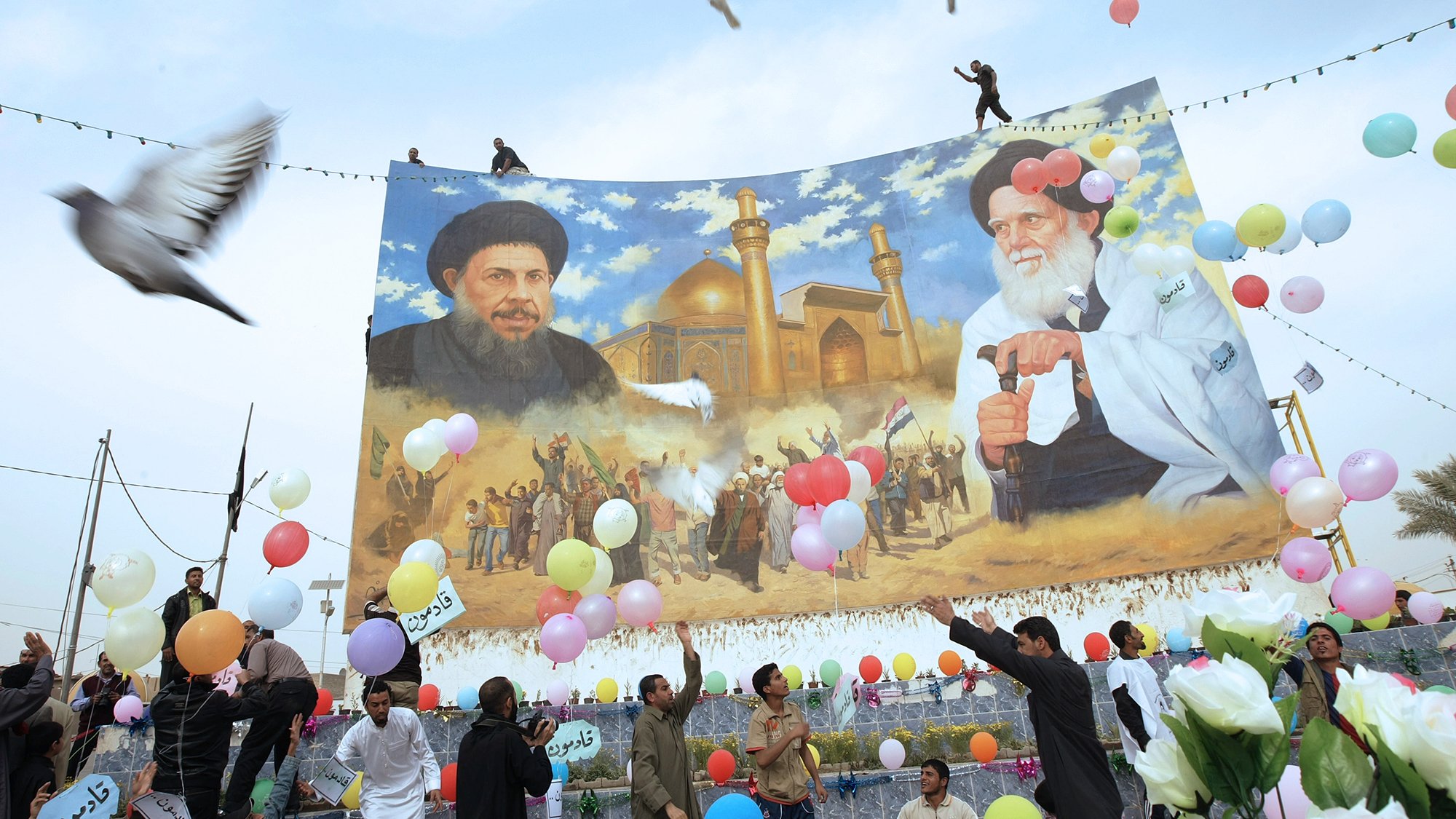 تظهر الصورة احتفالًا جماهيريًا حيث يحتشد مجموعة من الأشخاص تحت لافتة كبيرة تحتوي على صور لشخصيتين بارزتين. يبدو أنهم يقومون بإطلاق بالونات ملونة، ومن حولهم أجواء من الفرح والاحتفالات. في الخلفية، يُمكن رؤية معلم معماري يُحتمل أن يكون مسجدًا أو مكانًا مقدسًا. الألوان الساطعة والأجواء الحيوية تعكس روح الاحتفال والوحدة في المجتمع.