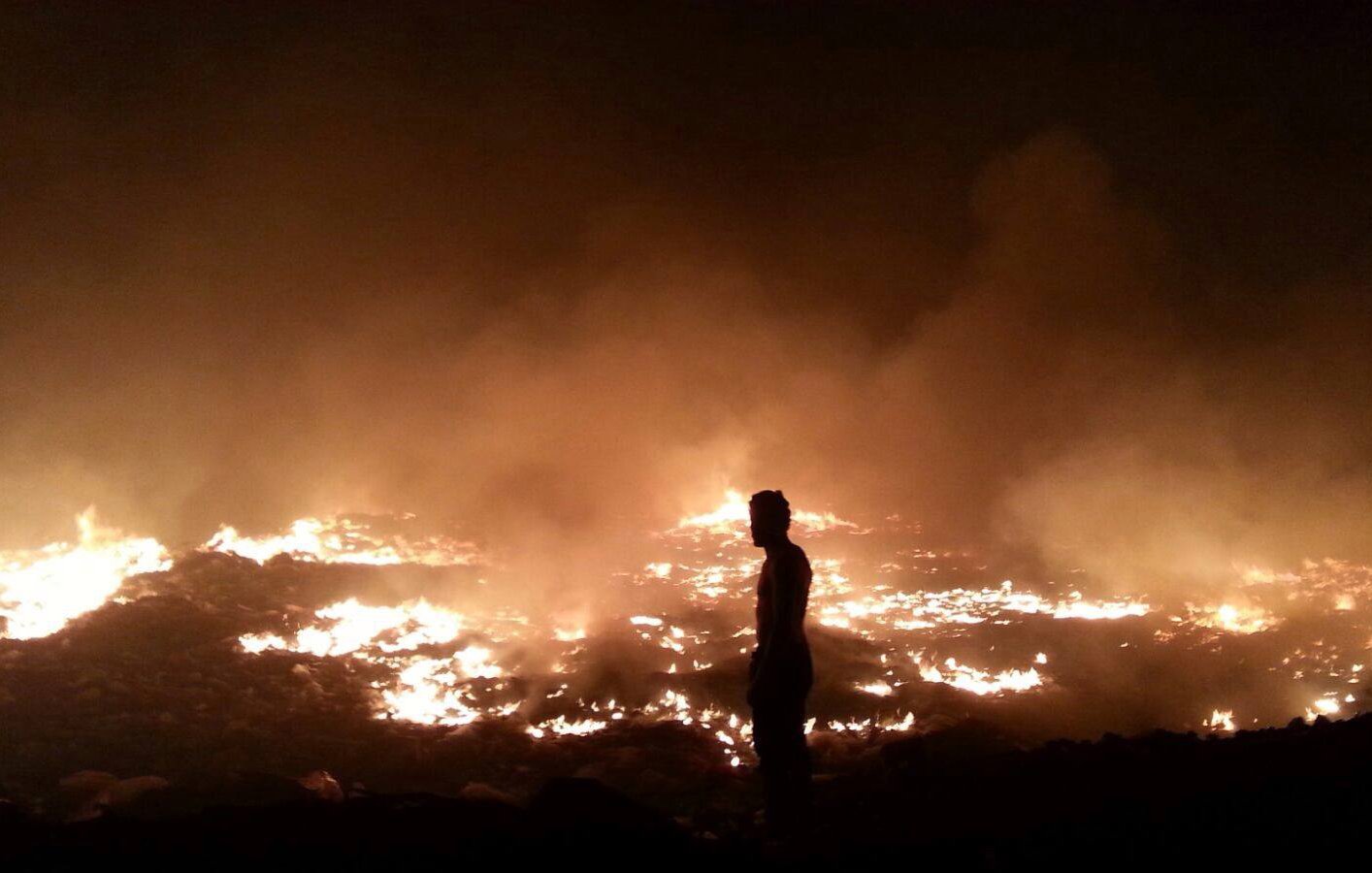 L'image montre un paysage de feu et de fumée. On voit un homme de silhouette, se tenant debout au premier plan, face à un vaste champ d'incendie. Le sol est enflammé, et une épaisse fumée s'élève, créant une atmosphère sombre et chaotique. Les flammes et la lumière orangée contrastent avec l'obscurité environnante, soulignant la gravité de la situation.