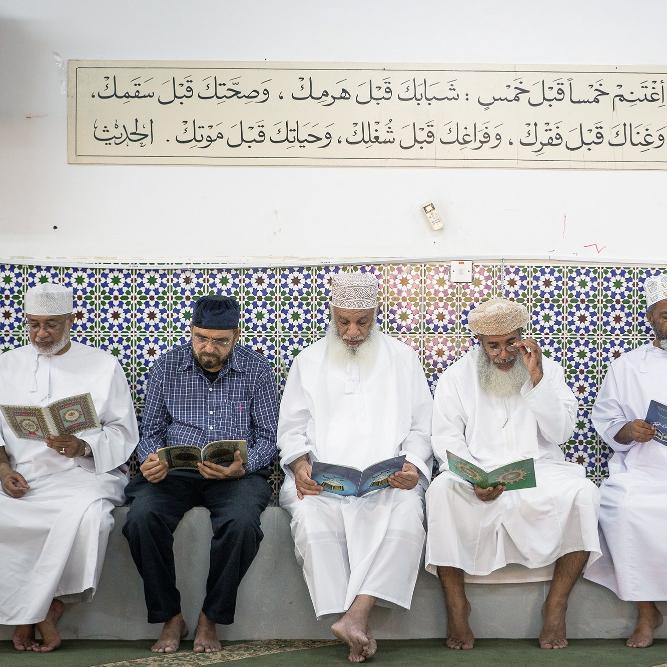 تظهر في الصورة مجموعة من الرجال يجلسون في صف واحد داخل مكان يبدوا أنه مسجد أو قاعة صلاة. كل منهم يحمل كتاباً، ويبدو أنهم يقرؤون أو يتدارسون. الجدران مزينة ببلاط ملون مزخرف، وهناك لوحة نصية مثبتة في الأعلى تحتوي على آيات أو نصوص دينية. يظهر في الصورة أجواء من التركيز والسكينة، مع تعبيرات الهدوء على وجوههم.