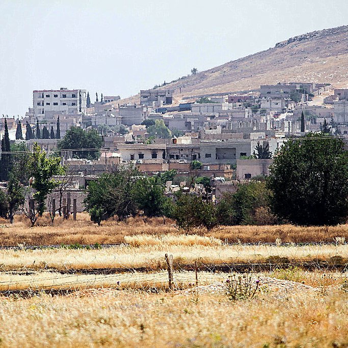 تظهر الصورة منطقة ريفية حيث يتواجد عدد من المنازل والمباني على تلة. في الخلفية، يمكن رؤية جبال منخفضة، بينما في المقدمة يوجد حقل جاف بألوان ذهبية. تبرز بعض الأشجار والنباتات هنا وهناك، مما يضفي لمسة من الخضرة على المشهد. كما يوجد خزان مياه في الجانب الأيمن من الصورة، مما يشير إلى سكان المنطقة. الجو يبدو مشمسًا مع أجواء صيفية.