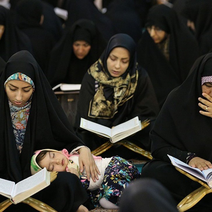 L'image montre un groupe de femmes, toutes vêtues de vêtements sombres, assises en train de lire des livres, vraisemblablement des copies du Coran. Certaines femmes sont profondément concentrées sur leur lecture, tandis qu'une d'elles tient un enfant qui semble s'endormir sur ses genoux. L'atmosphère est sérieuse et contemplative, indiquant un moment de prière ou de méditation collective. Les femmes portent des hijabs, ce qui est caractéristique de certaines traditions culturelles et religieuses. L'environnement semble être un espace communautaire, peut-être dans une mosquée ou un lieu de réunion.