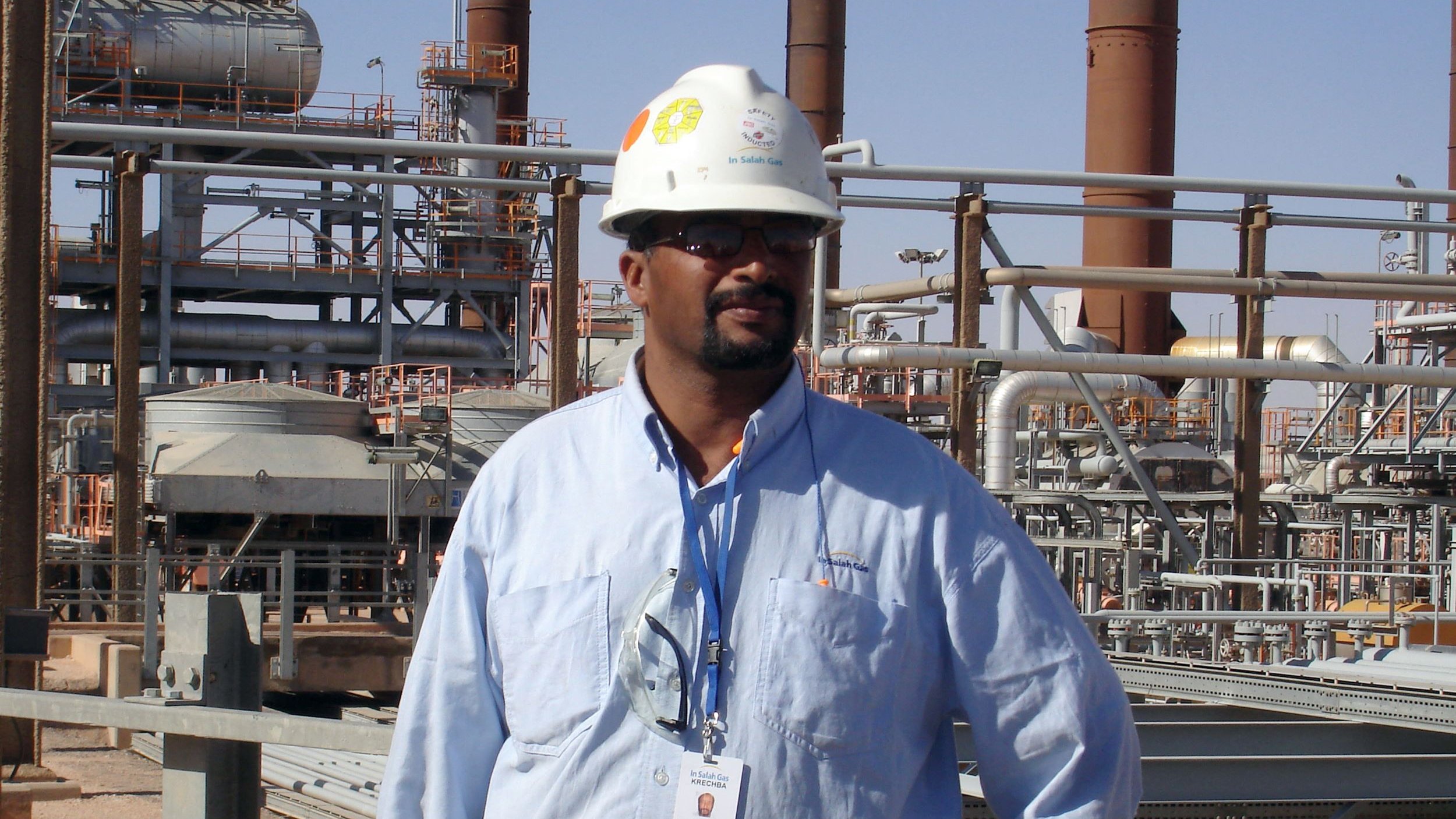 L'image montre un homme portant un casque de sécurité blanc et une chemise bleue, debout dans un environnement industriel. En arrière-plan, on aperçoit des structures métalliques et des installations, probablement liées à une usine ou une raffinerie. L'homme semble professionnel et attentif, avec une carte d'identification suspendue à son cou.